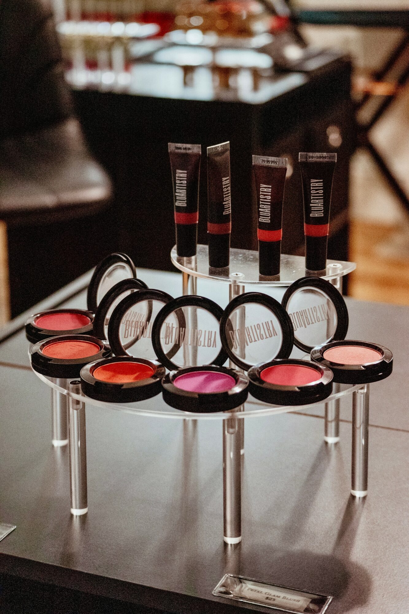 Display of lip glosses and blushes on a round, multi-tiered stand in a store setting.