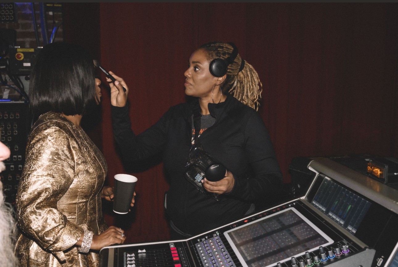 Two women in a recording studio, one applying makeup or adjusting the other's makeup, with audio equipment nearby.