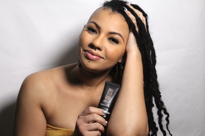Woman with dreadlocks and a yellow strapless top, smiling and holding a small black object, against a plain background.