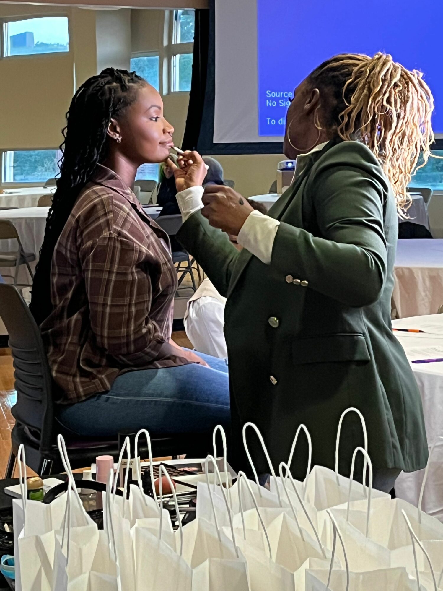 Two women engaging in a conversation indoors, with a table of gift bags nearby, and a screen in the background.
