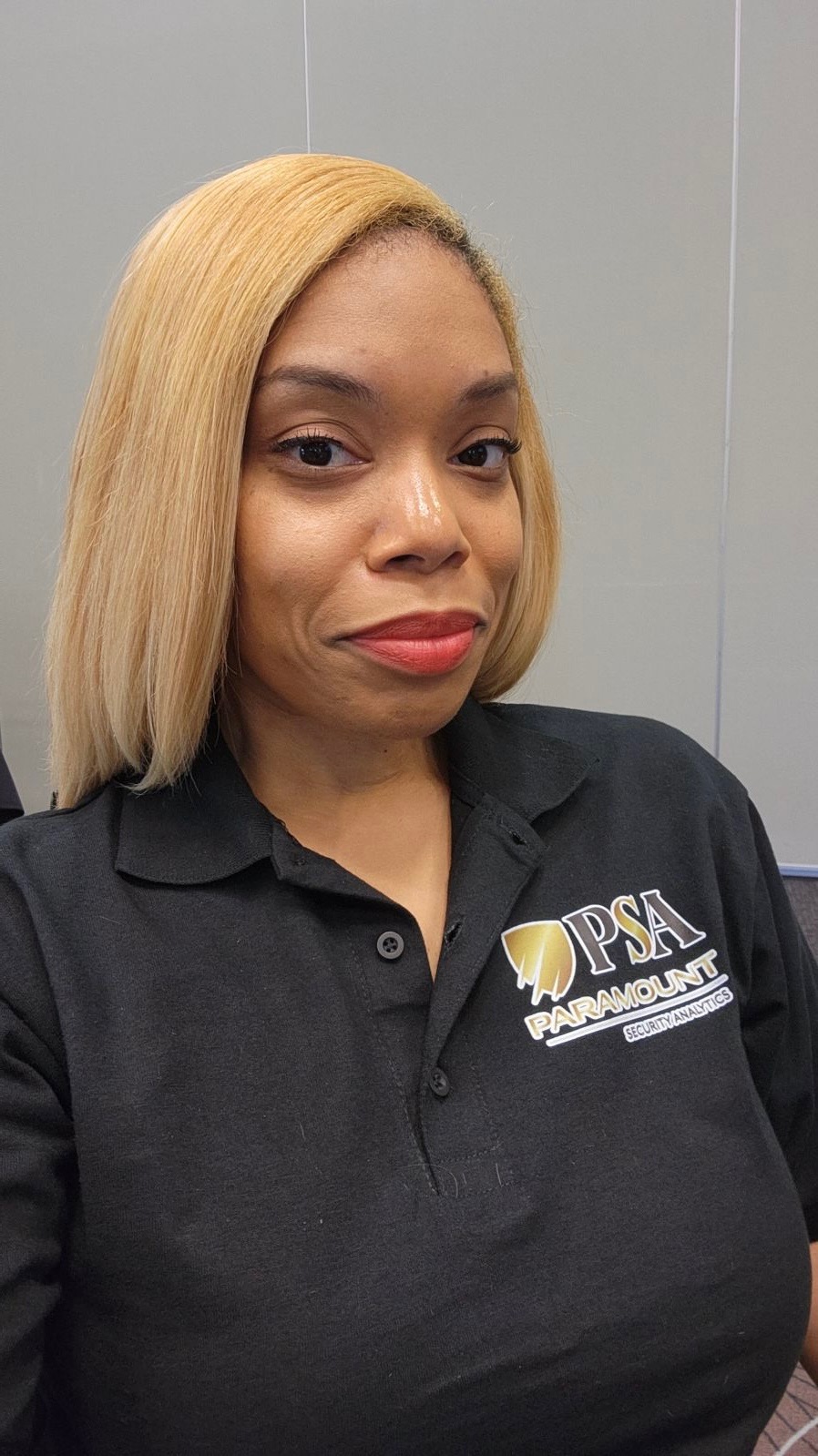 Woman with blonde hair wearing a black polo shirt with a logo, standing against a plain background.