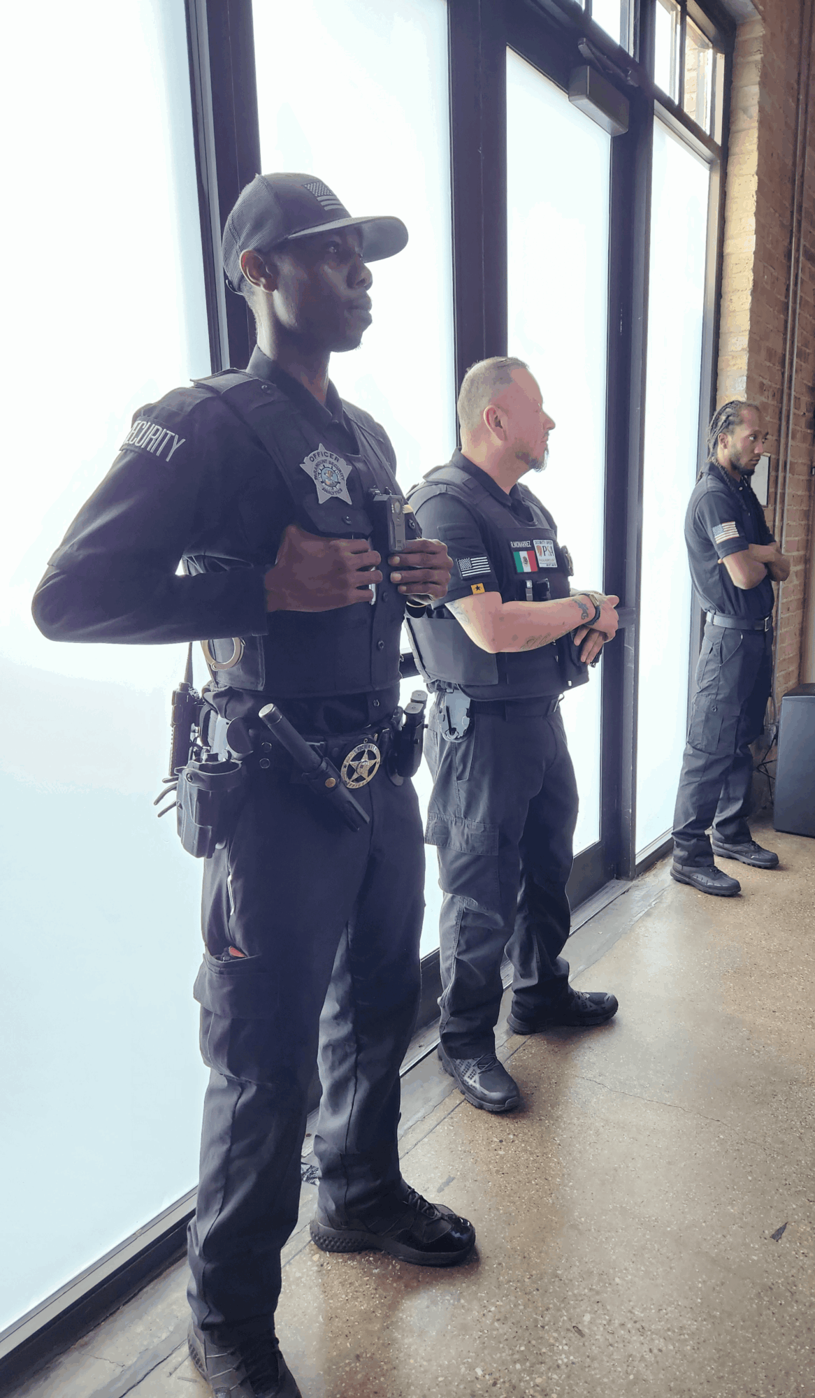 Three police officers standing indoors near large windows, facing sideways, with hands clasped or at their sides.
