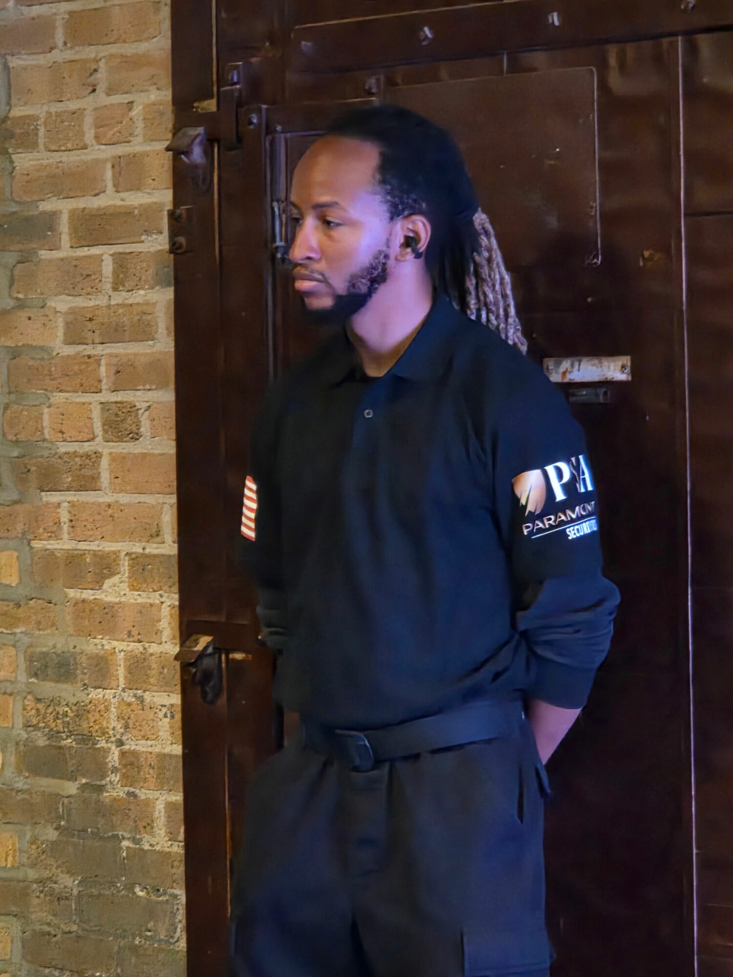 Person with long braided hair leaning against a wooden surface, wearing a dark shirt and pants, with a brick wall background.