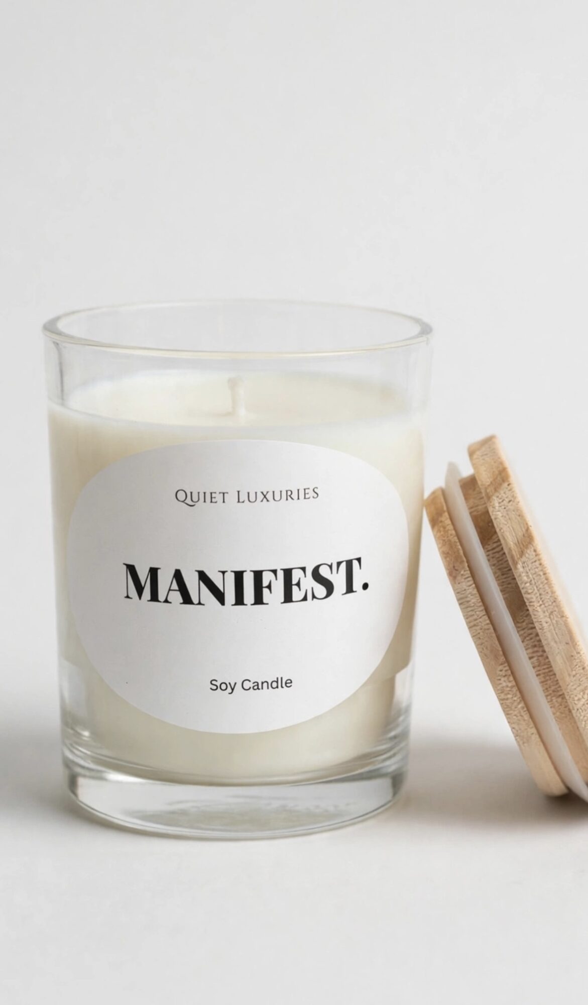 Soy candle in a glass container with a wooden lid beside it, label reads 'MANIFEST.' and 'Quiet Luxuries'.