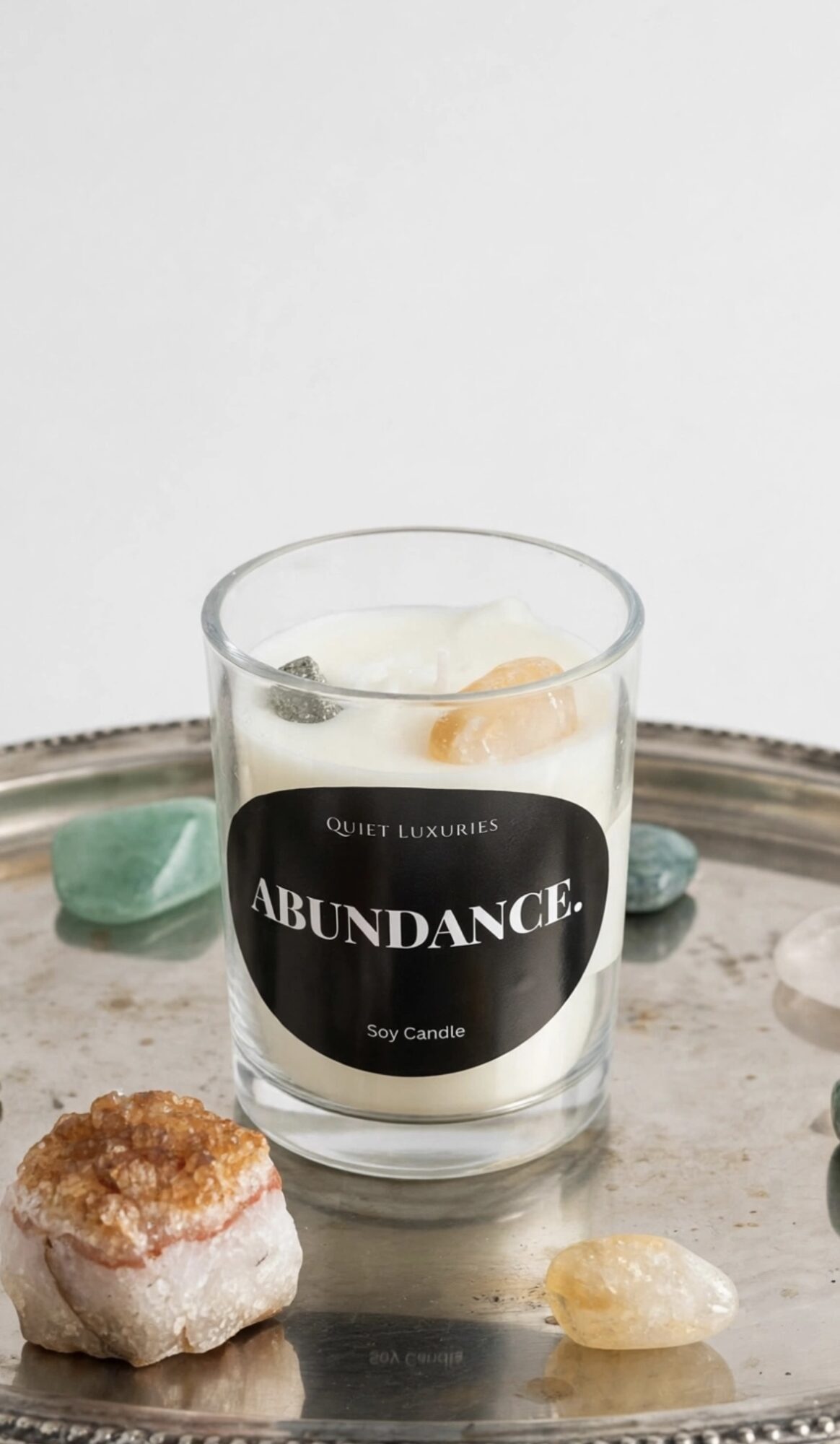 A candle in a glass container labeled 'ABUNDANCE' on a tray with decorative stones, against a plain background.
