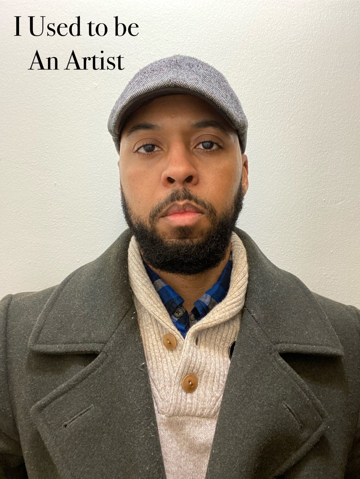 A man with a beard wearing a gray cap, coat, and sweater, standing against a plain wall, with text in the top left corner.