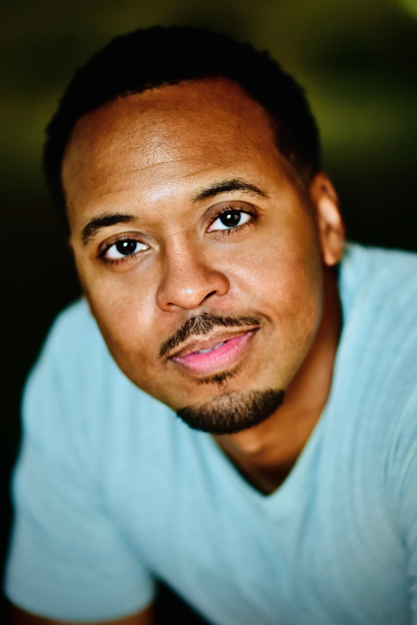 Close-up of a man with short curly hair and a goatee, wearing a light blue shirt, smiling softly.