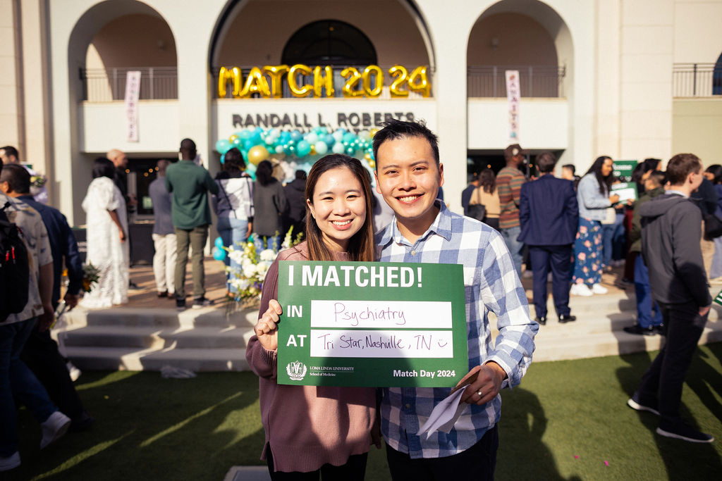 Two smiling people holding a green sign with white text at a graduation event outside a building with a yellow sign reading 'MATCH 2024'.