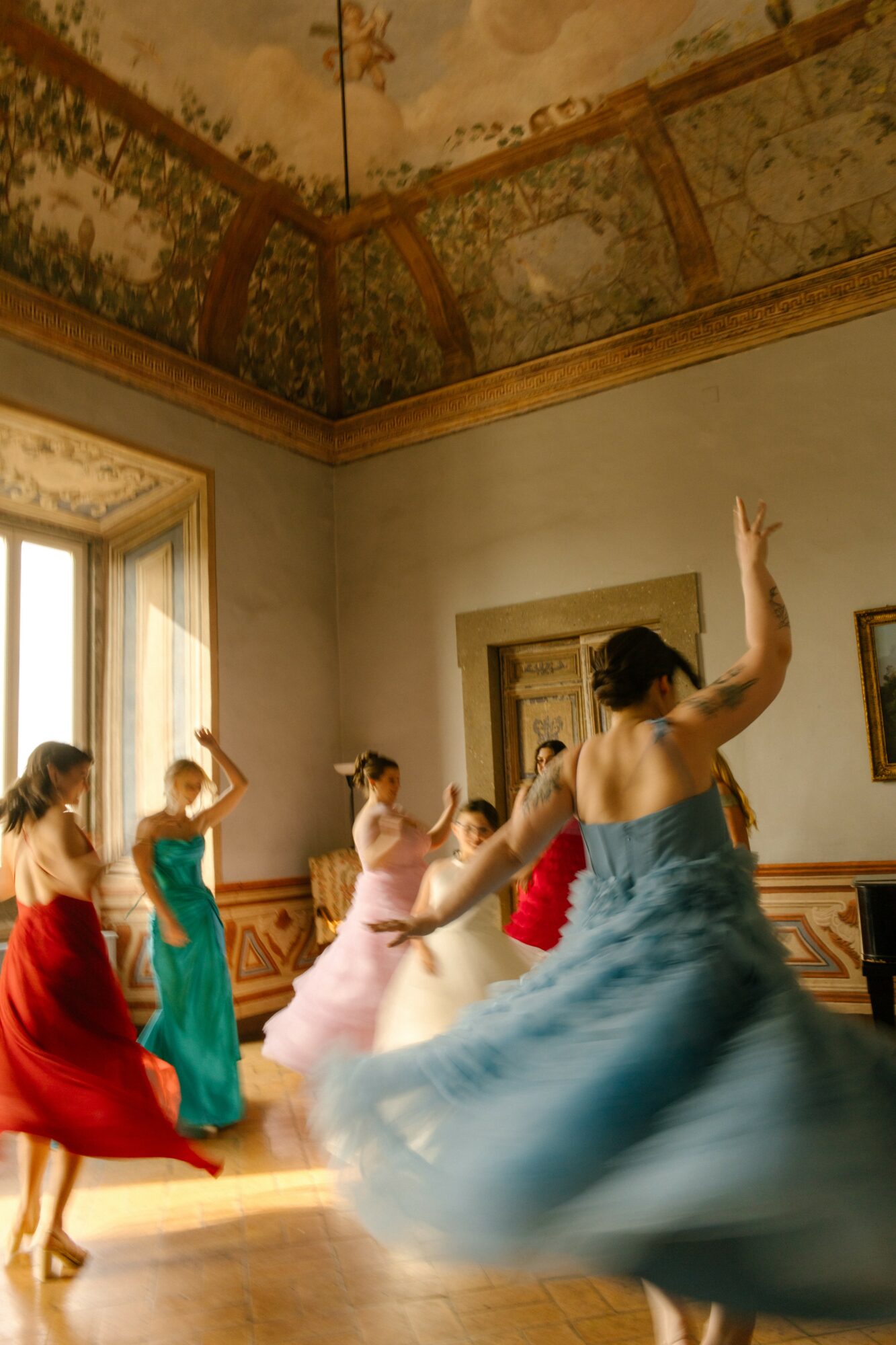 Group of women dancing in a room with ornate ceiling and sunlight through window, wearing colorful dresses.