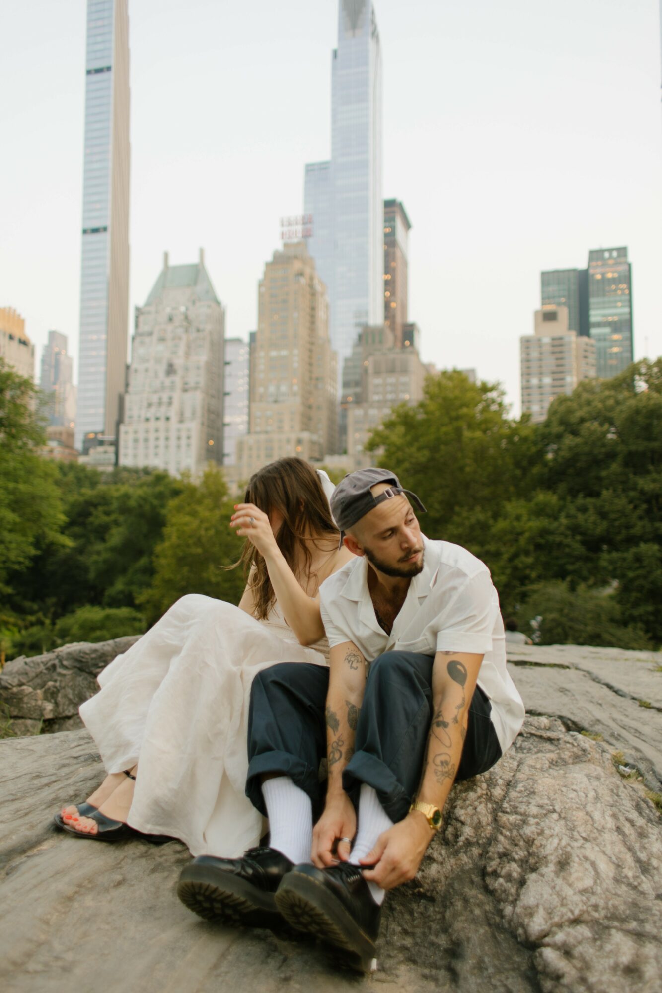 A couple sitting on a rock in a park with city skyscrapers in the background, the woman in a white dress and the man in casual clothing.