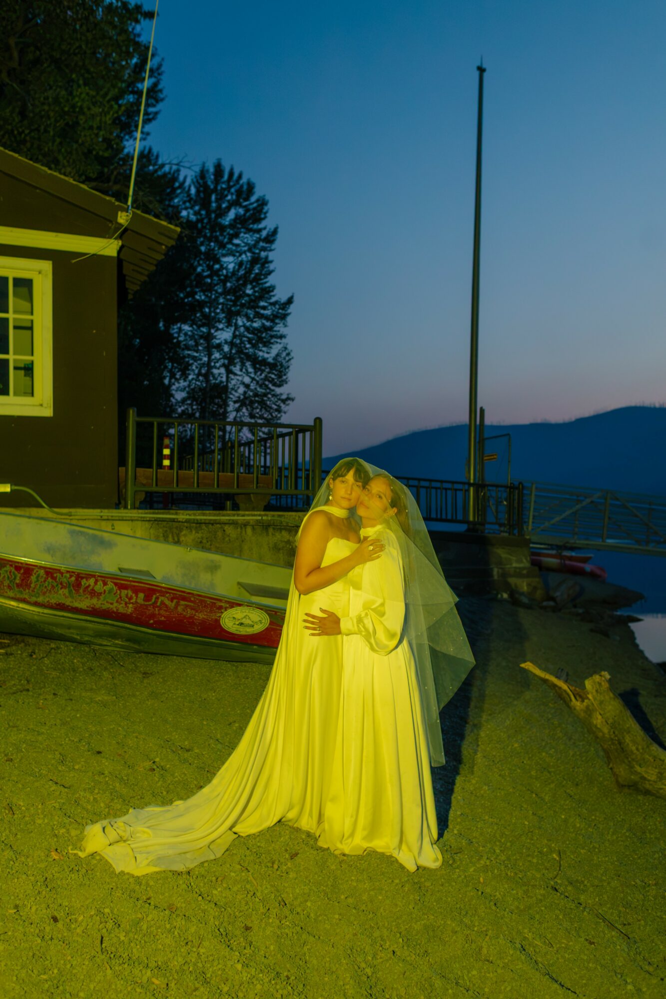 Person in a long yellow dress with a veil, standing outdoors near water, with a building and trees in the background.