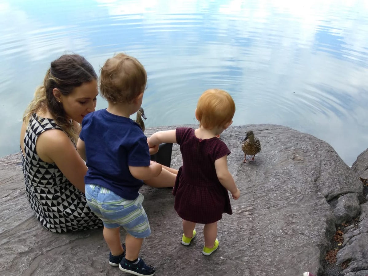 Three children and a duck on a large rock by water, with the children watching the duck.
