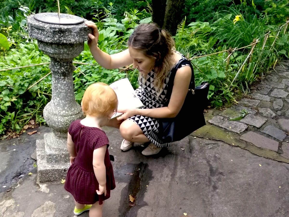 Young woman sitting on stone pavement, reading a book to a small girl near a garden fountain, surrounded by greenery.