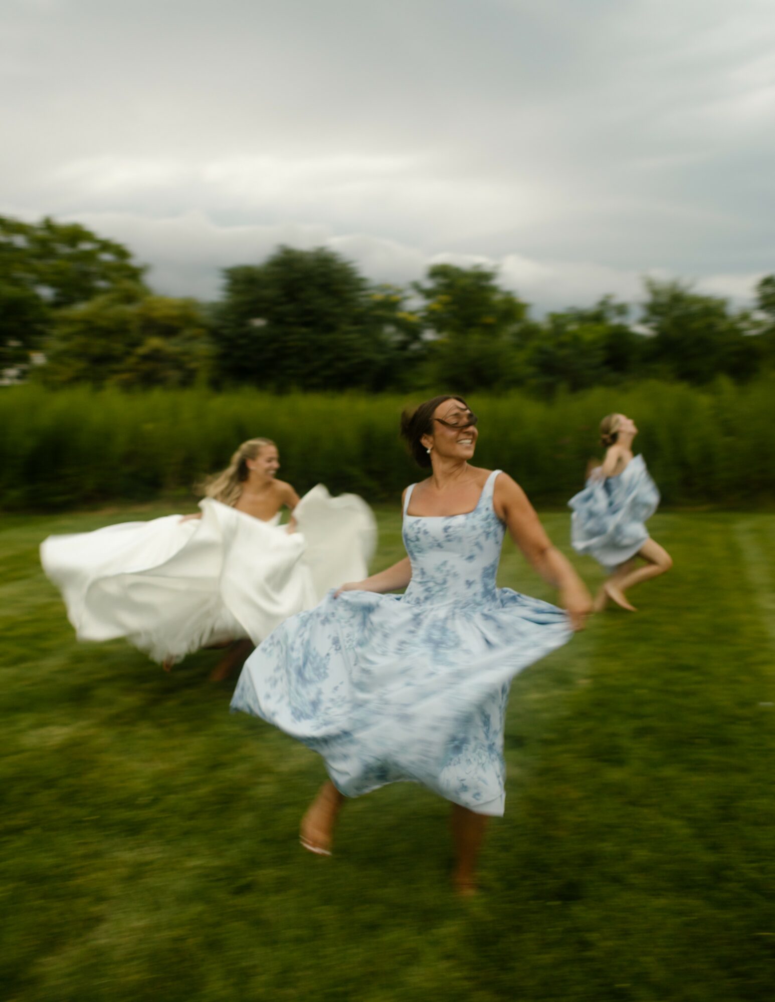 Three women in dresses running on grass with trees and cloudy sky in background.
