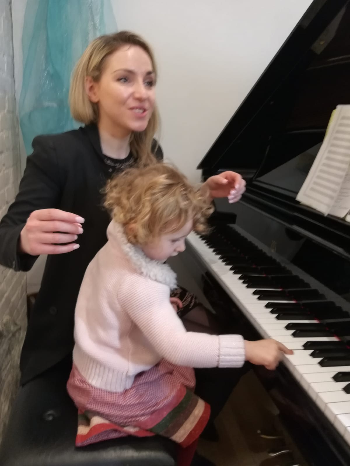 Woman and child playing piano together, woman smiling, child reaching for keys, indoor setting with a curtain in the background.