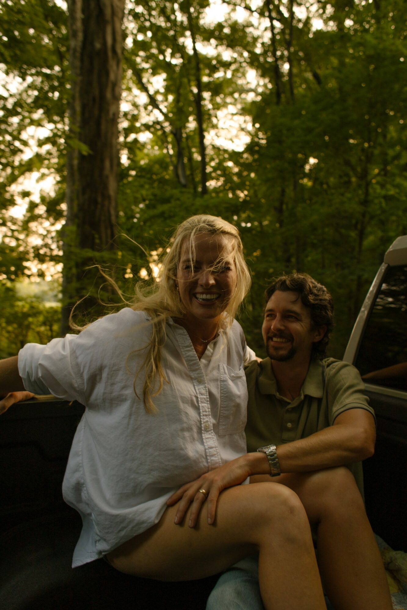 Two people sitting outdoors in a forest, smiling, with trees and sunlight in the background.