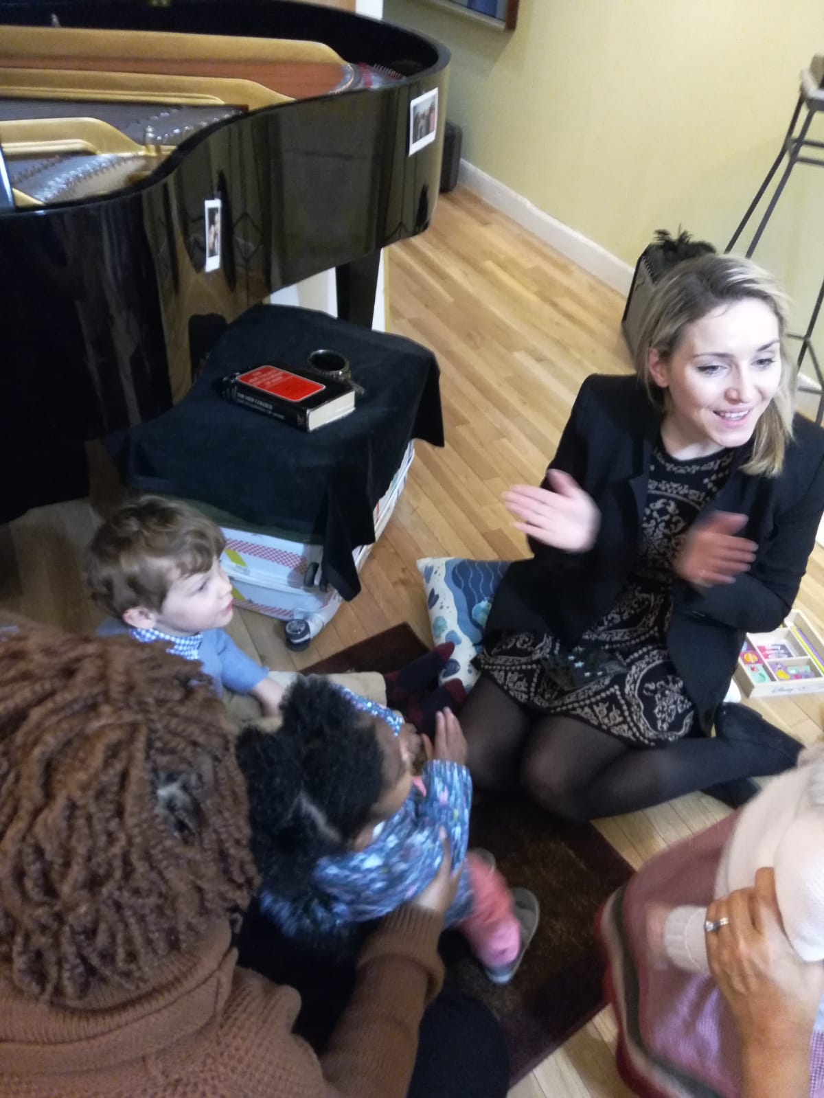 Young woman sitting on the floor with children, clapping and smiling, in a room with a piano and wooden floor.