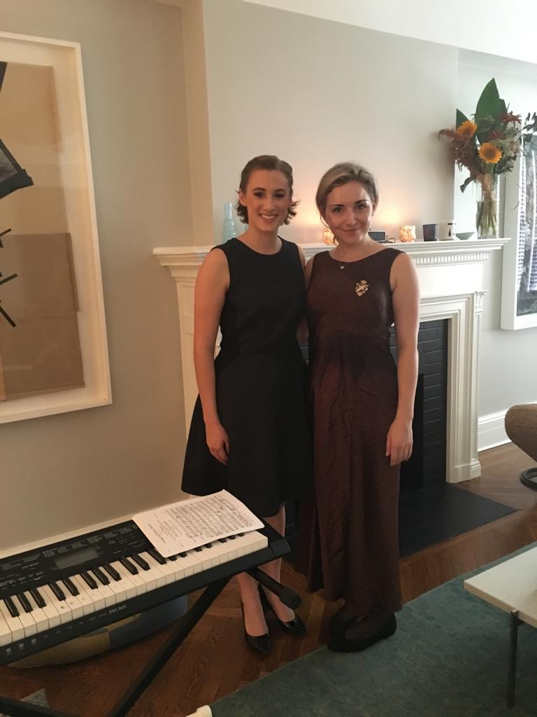 Two women stand side by side indoors, smiling, with a fireplace and floral arrangement in the background.