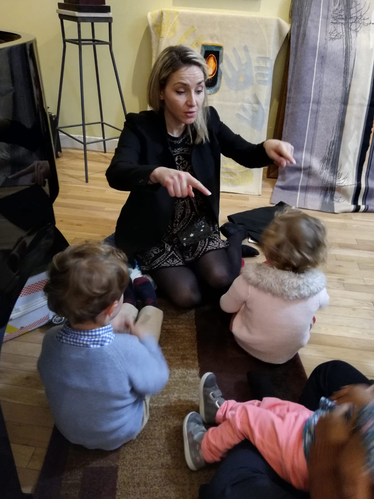 Woman sitting on floor with three children, engaging in conversation indoors, with furniture and curtains in background.