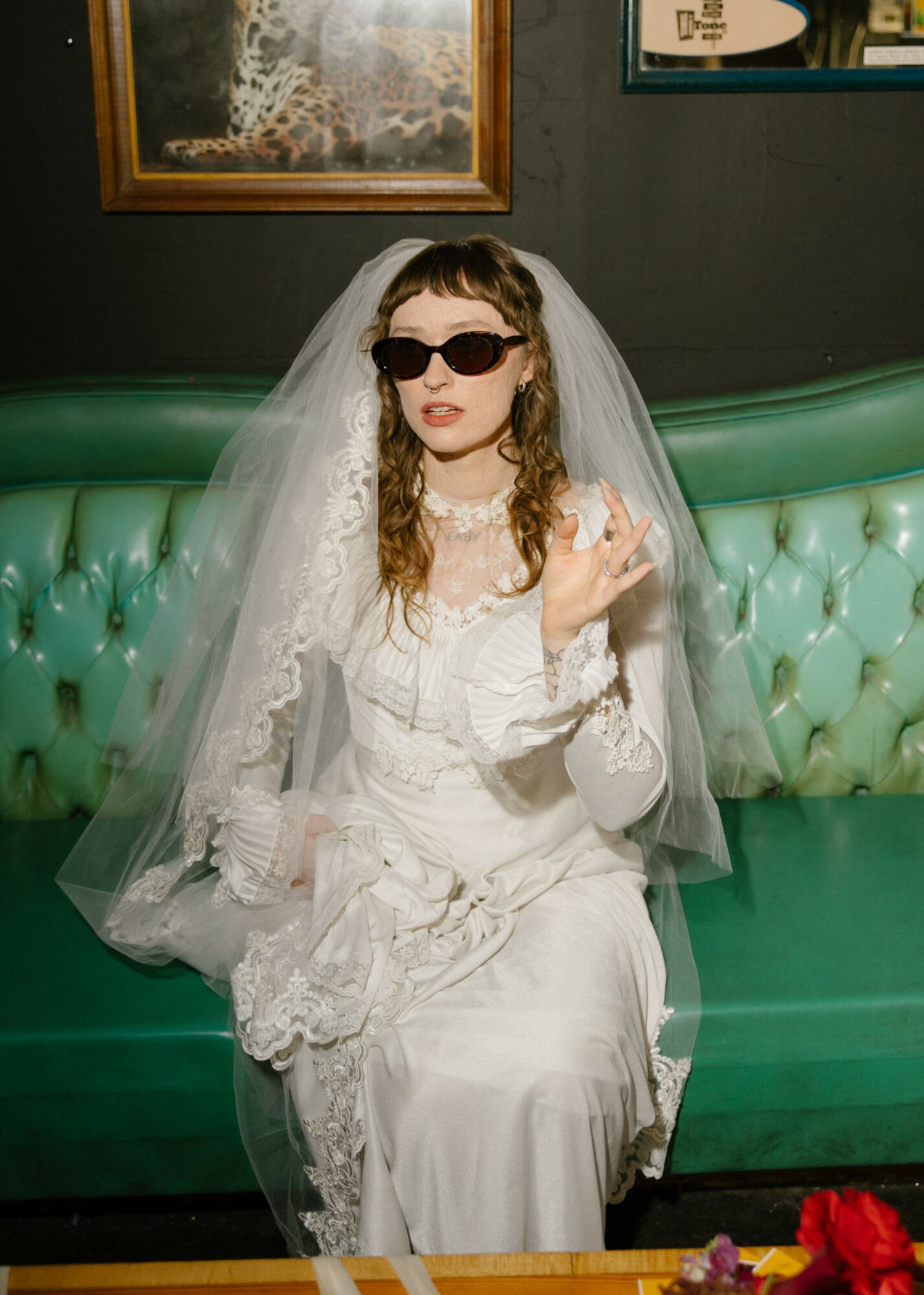 Woman in wedding dress with veil and sunglasses sitting on green sofa, holding a glass, in a room with framed artwork.