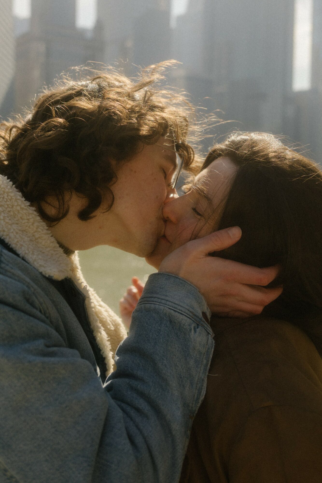 Two people kissing, one with curly hair and the other with straight hair, outdoors with city buildings in background.