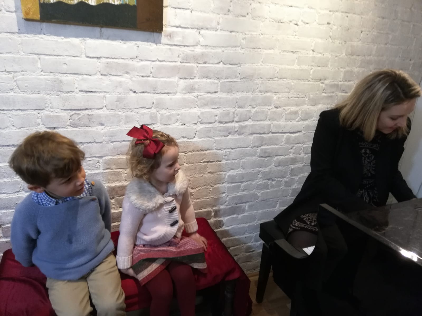 Two children sitting on a red cushion against a white brick wall, with a woman playing a piano nearby.