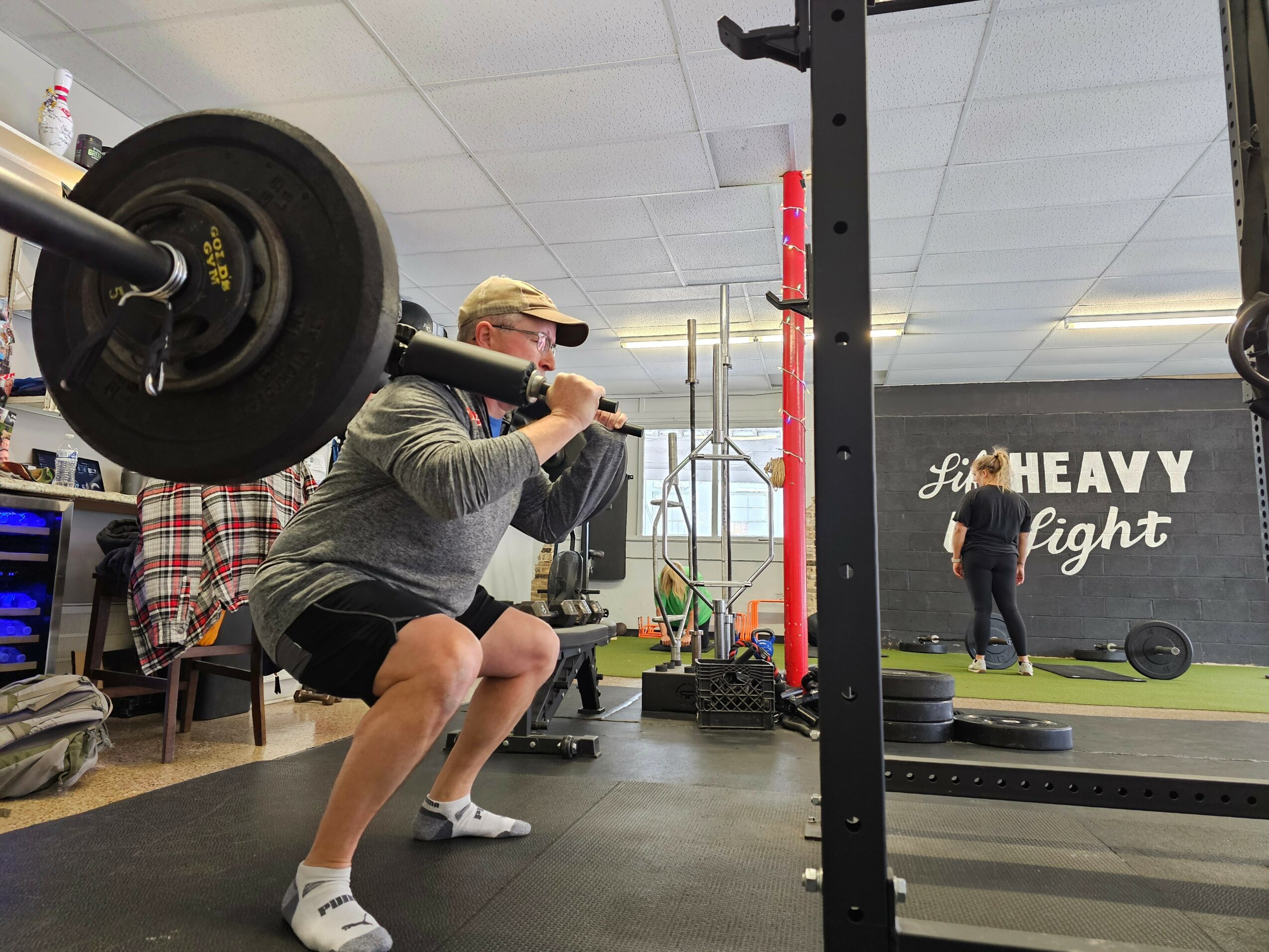 Person squatting and lifting a barbell in a gym, with another person in the background near a wall with gym equipment.