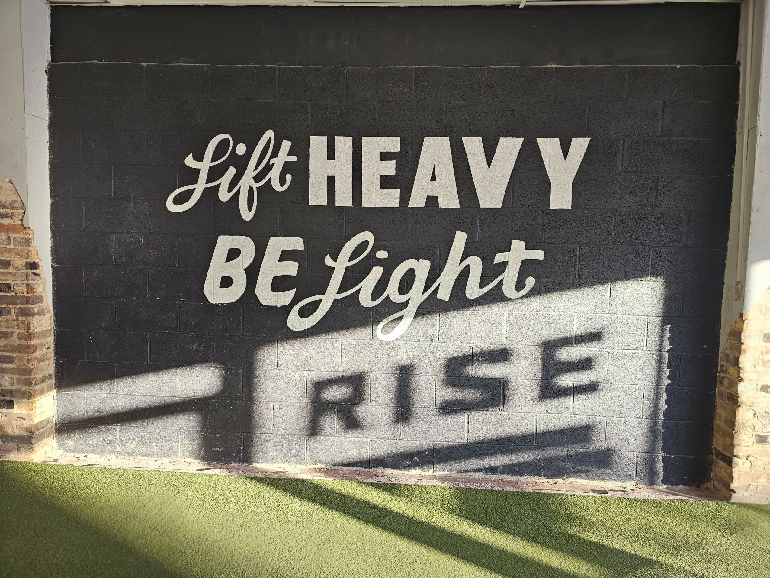 Wall with motivational quote: 'Lift HEAVY BE Light RISE' in white text on black background, shadowed by sunlight.