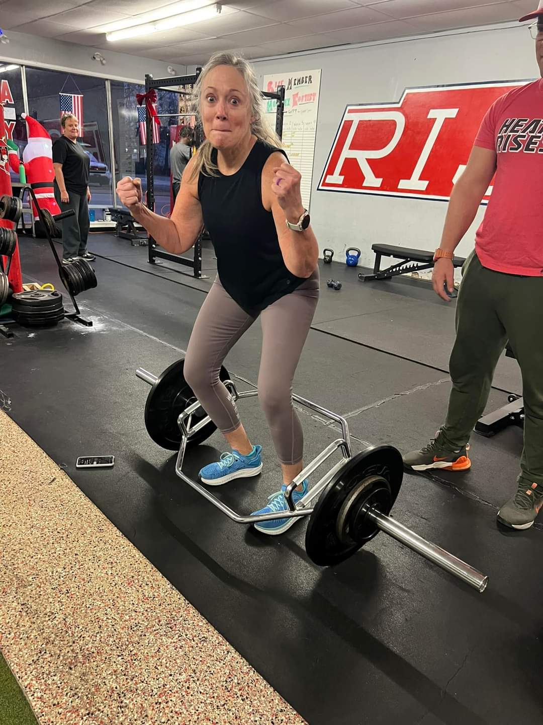 Woman in black top and gray leggings on a skateboard in a gym, with gym equipment and a person in red shirt nearby.