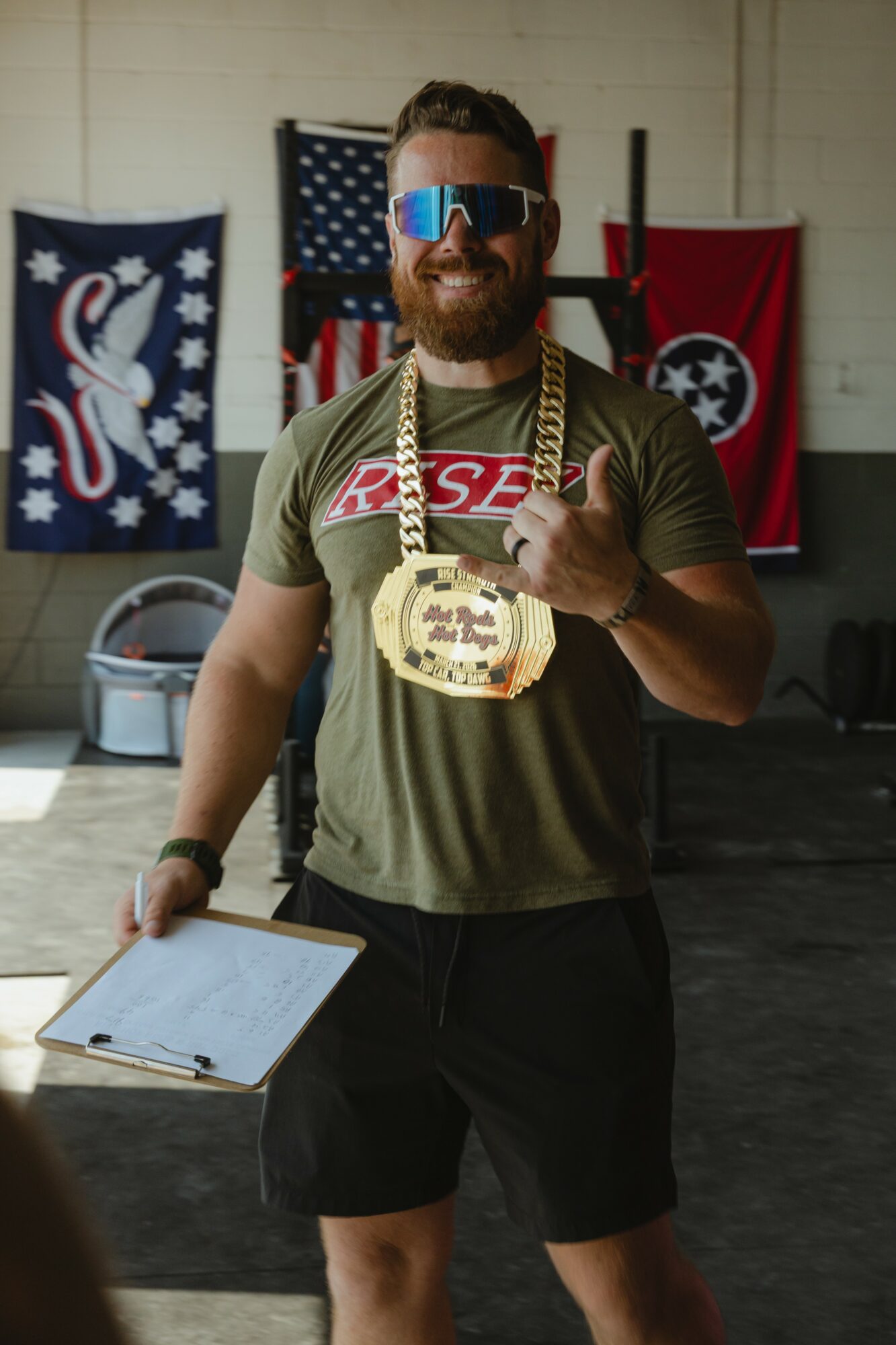 Man with sunglasses and a medal around his neck, holding a clipboard, smiling indoors with flags and banners in background.