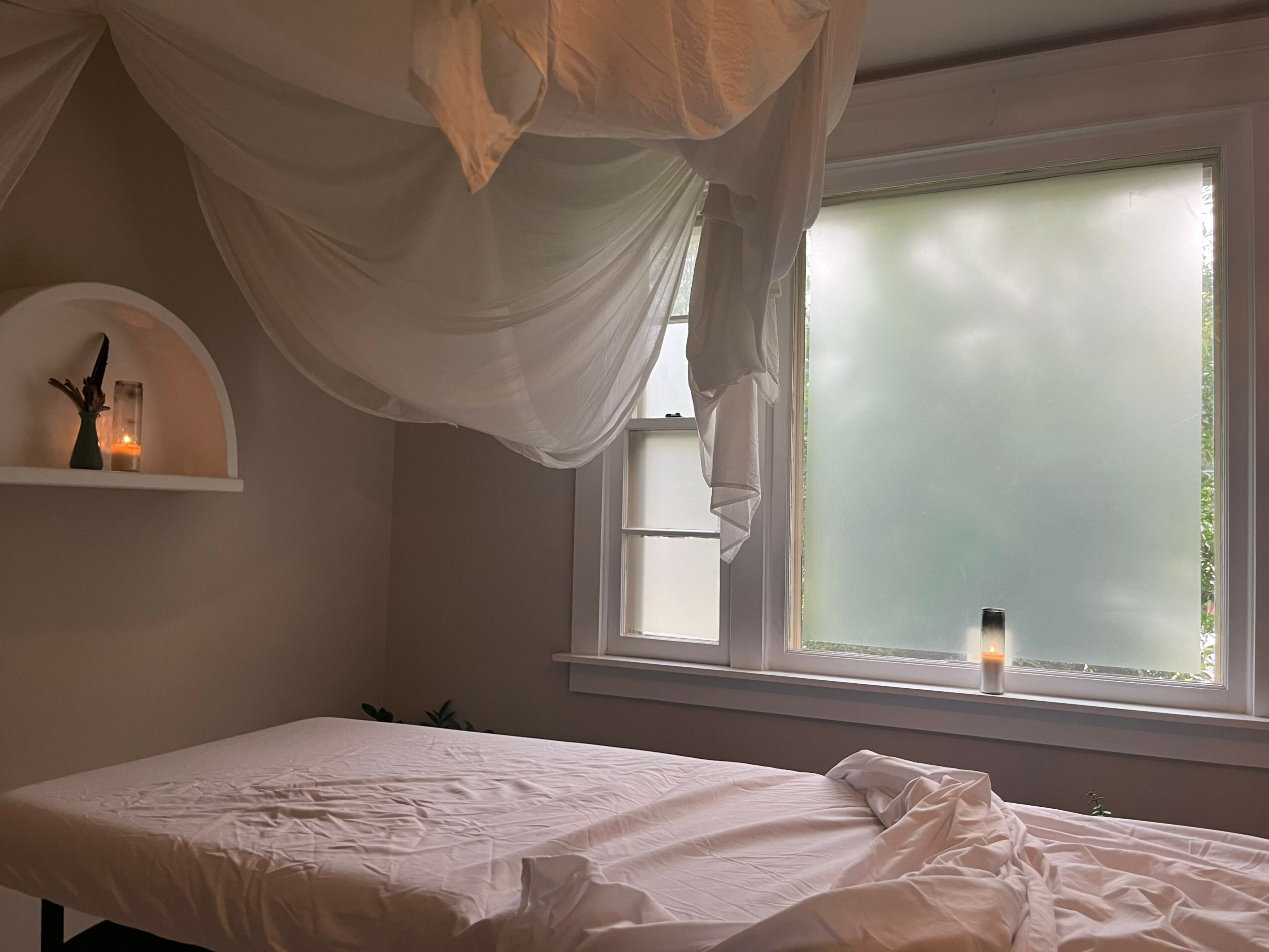 Unmade bed with a white canopy above, window with frosted glass, and a small candle on the windowsill.