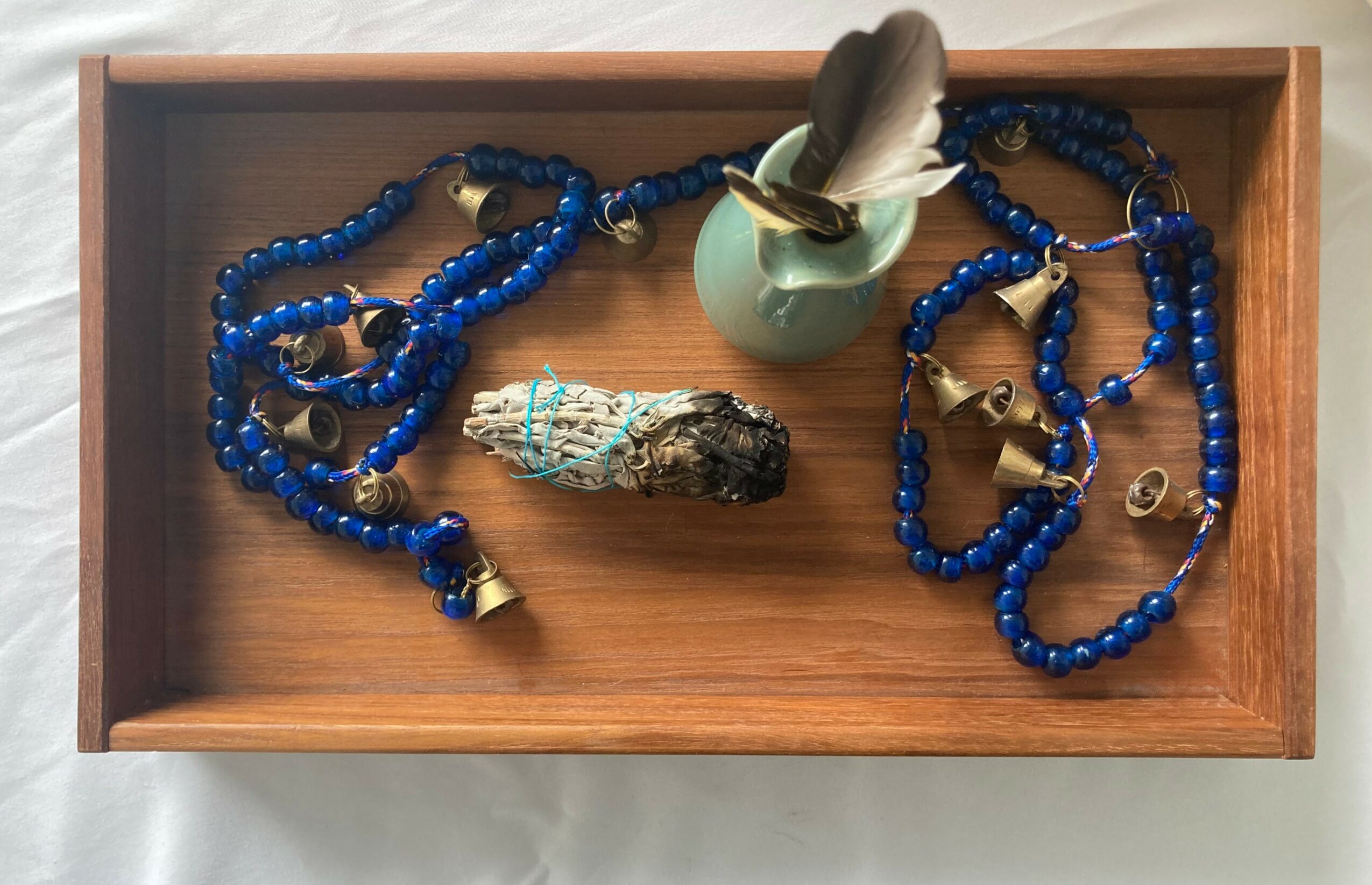 Wooden tray with blue beaded necklaces, a small ceramic cup with a feather, and a rock inside.