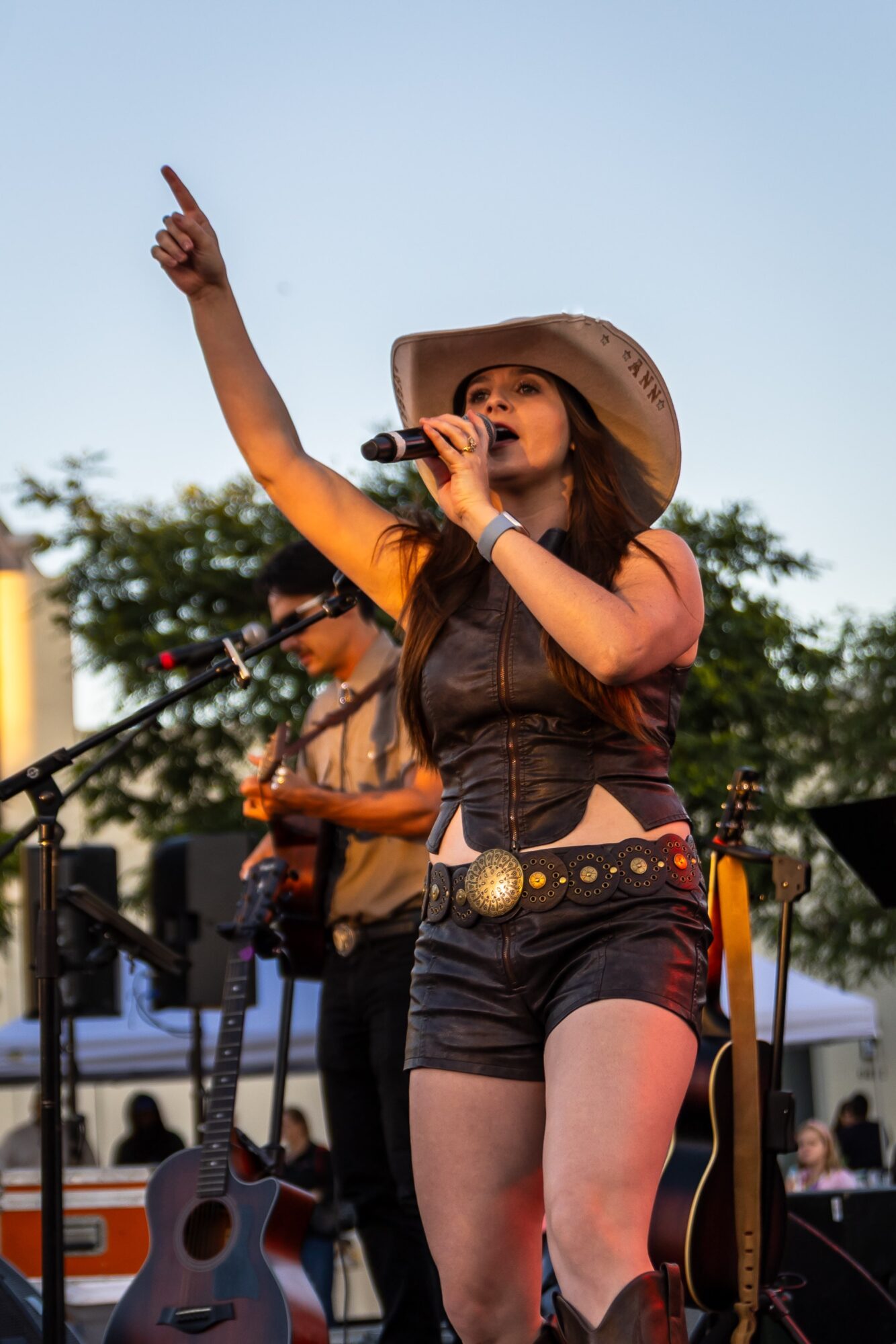 Female singer wearing a hat and black outfit performs outdoors, pointing upward with her right hand, holding a microphone in her left hand.