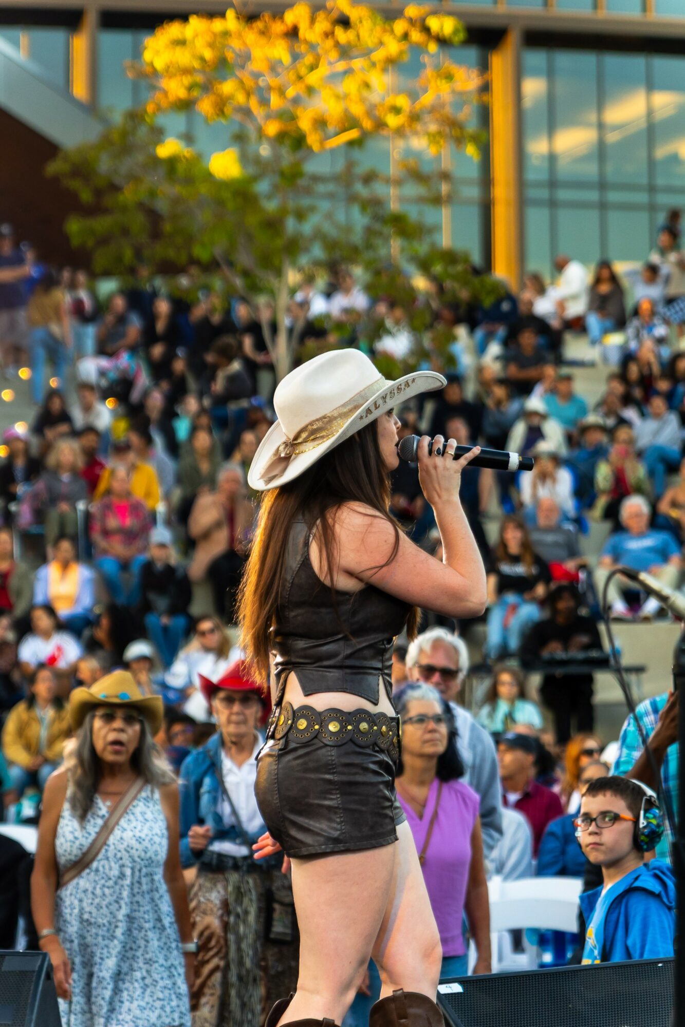 Woman with long hair wearing a large sun hat, black top, and shorts speaking into a microphone at an outdoor event, crowd in background.