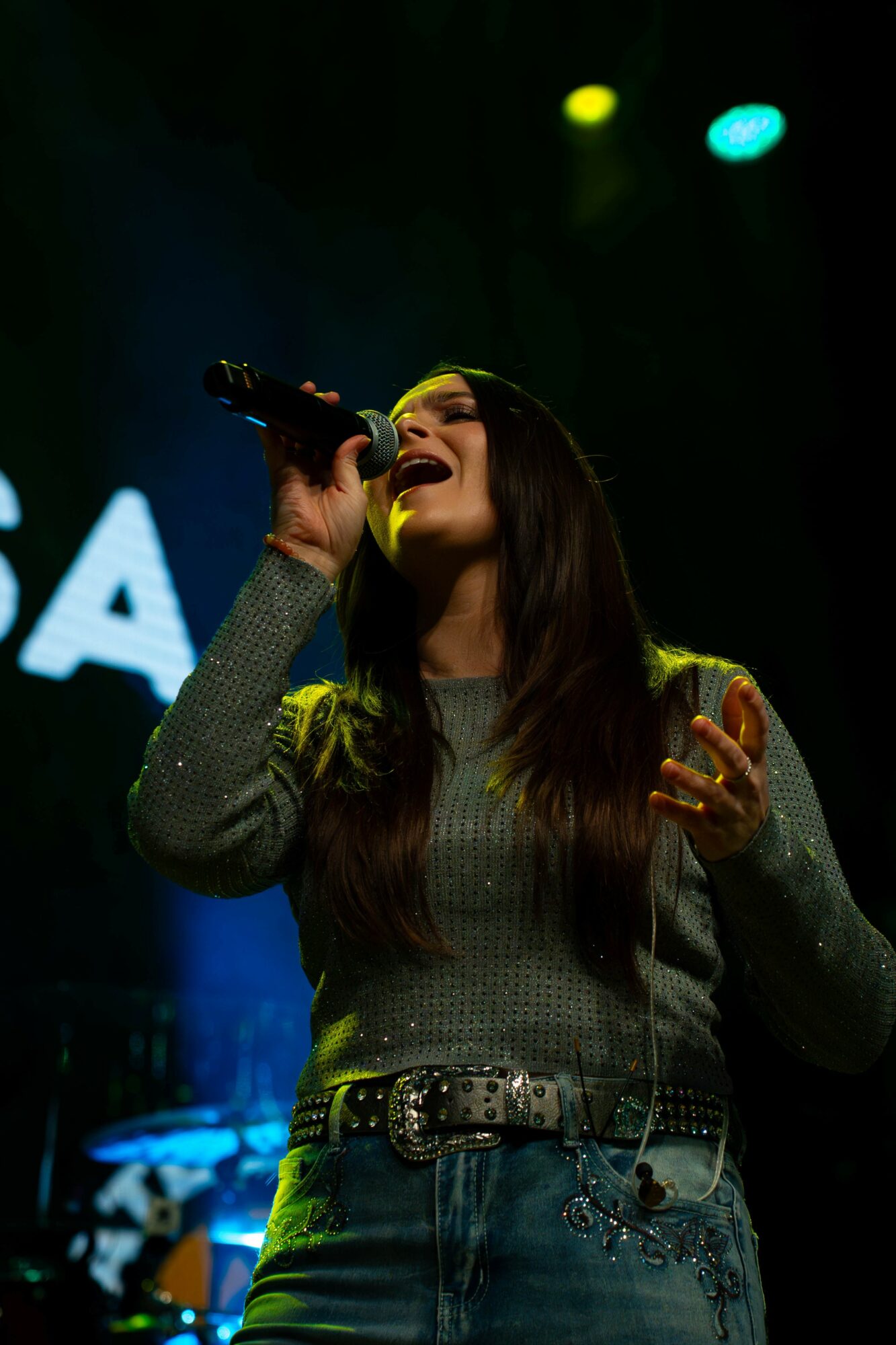 Woman singing into a microphone on stage with colorful lights, wearing a gray sweater and jeans.