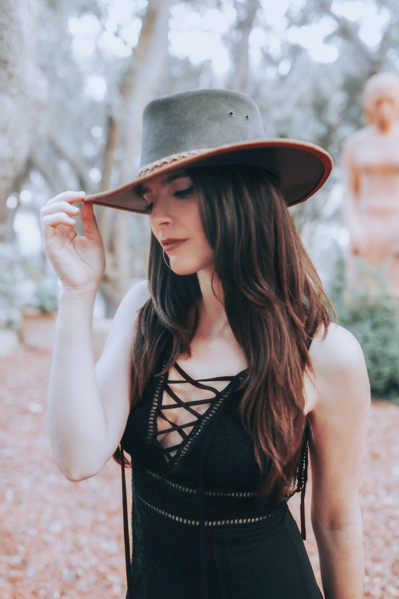 Woman wearing a wide-brimmed hat and black dress outdoors, looking down, with trees in the background.