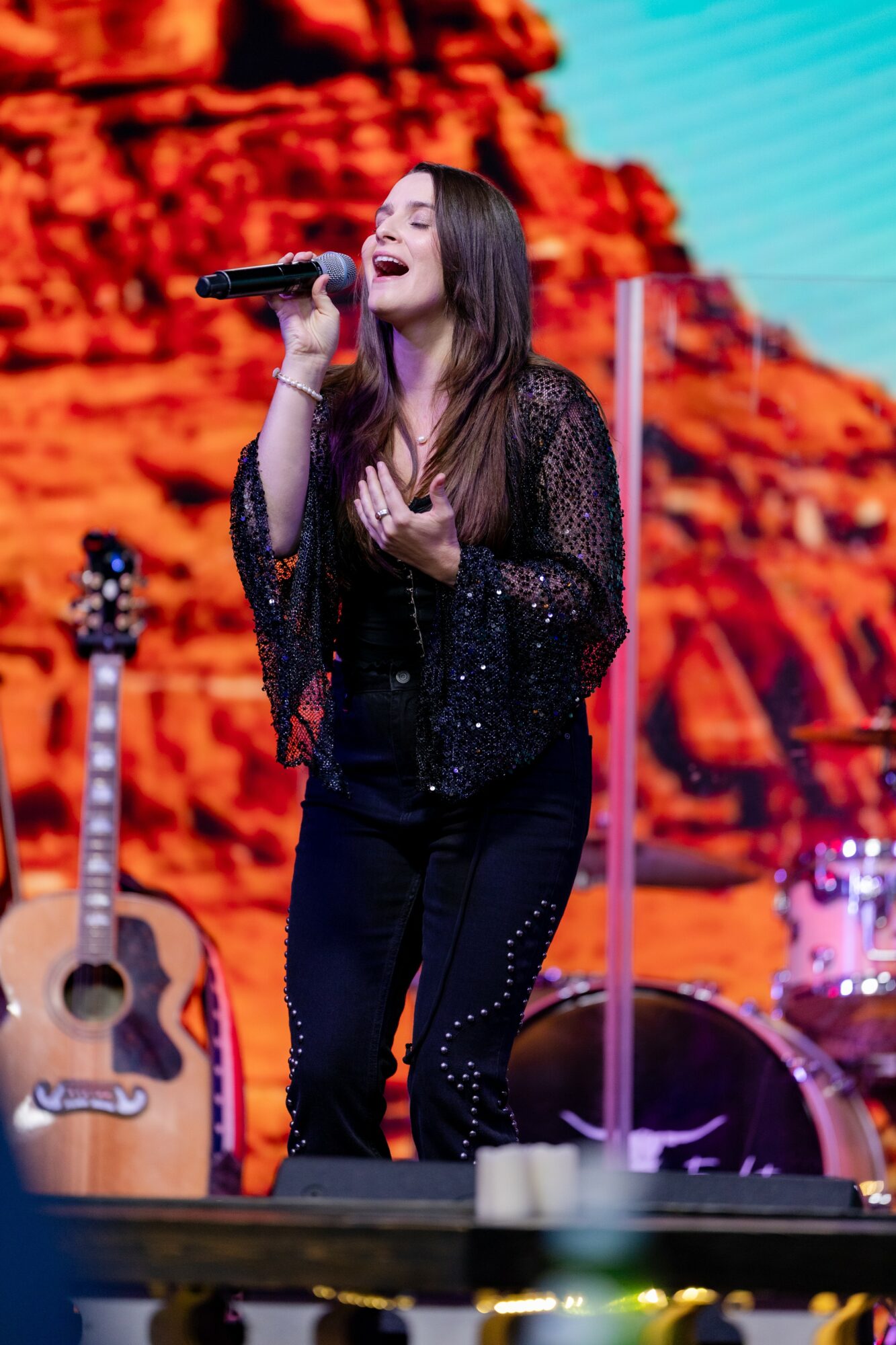 Female singer performing on stage with a desert landscape background, holding a microphone, wearing a black sparkly outfit.