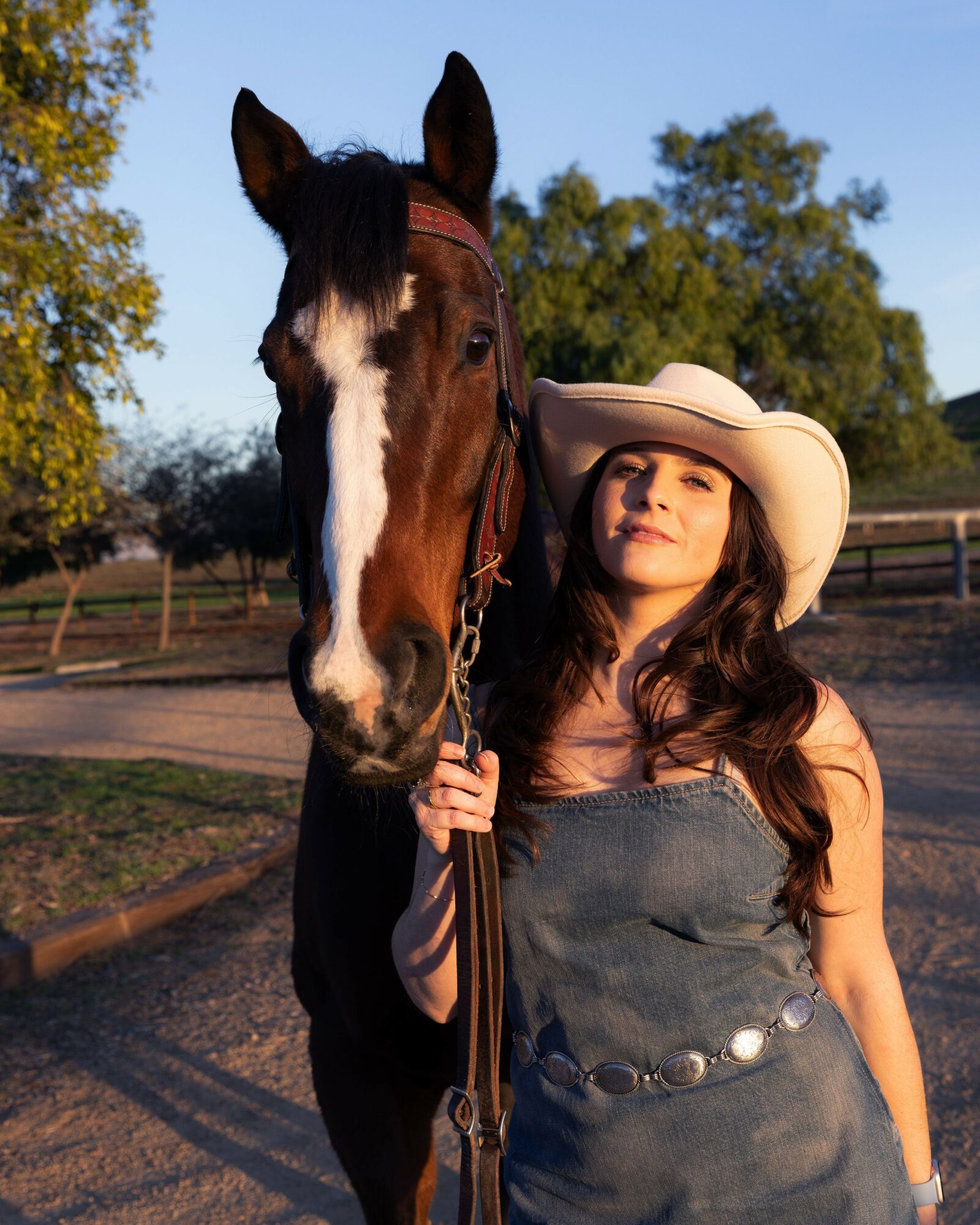 Woman in a wide-brimmed hat standing next to a horse outdoors, trees and a fence in the background.