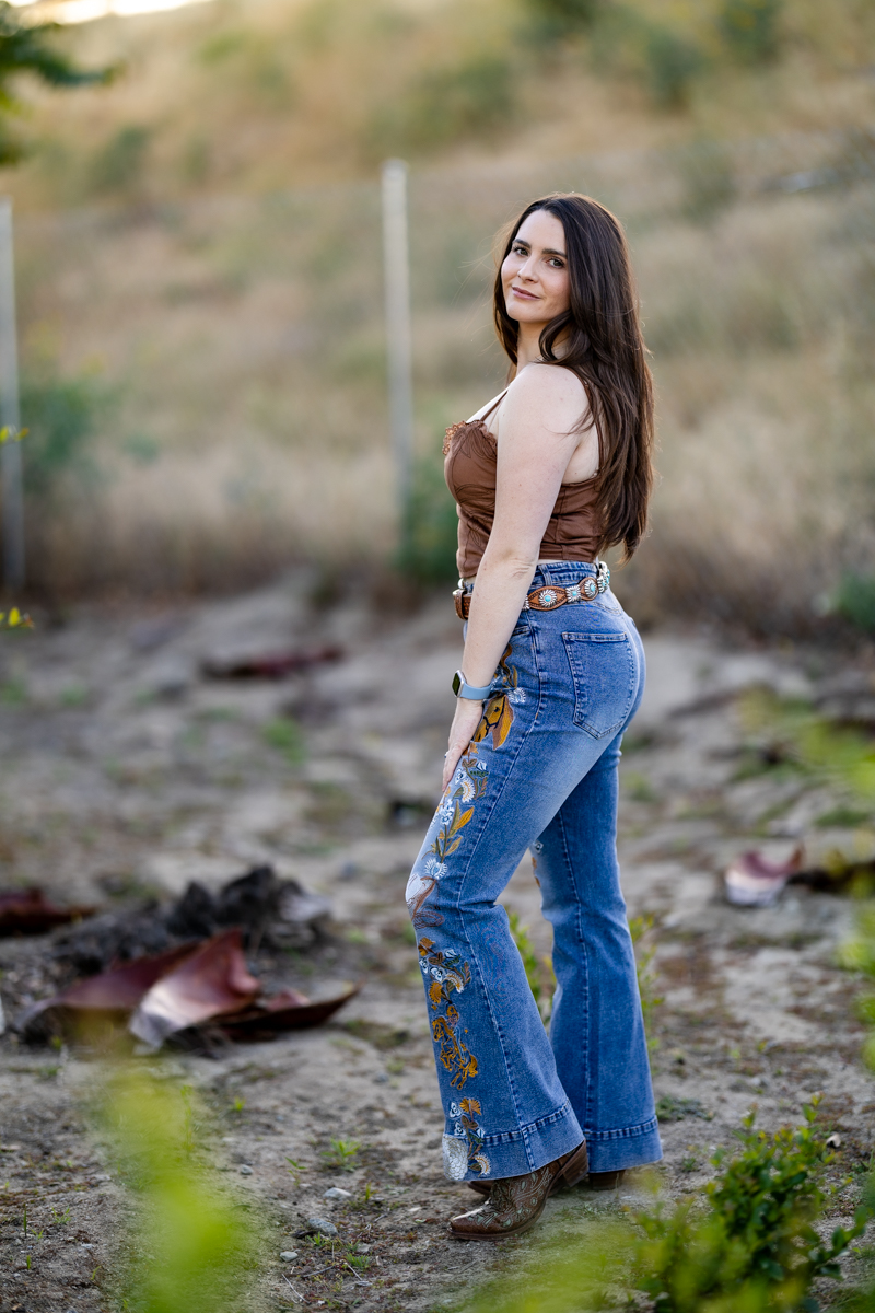 Young woman with long dark hair standing outdoors on a dirt path, wearing a beige top and blue jeans with floral embroidery, smiling.