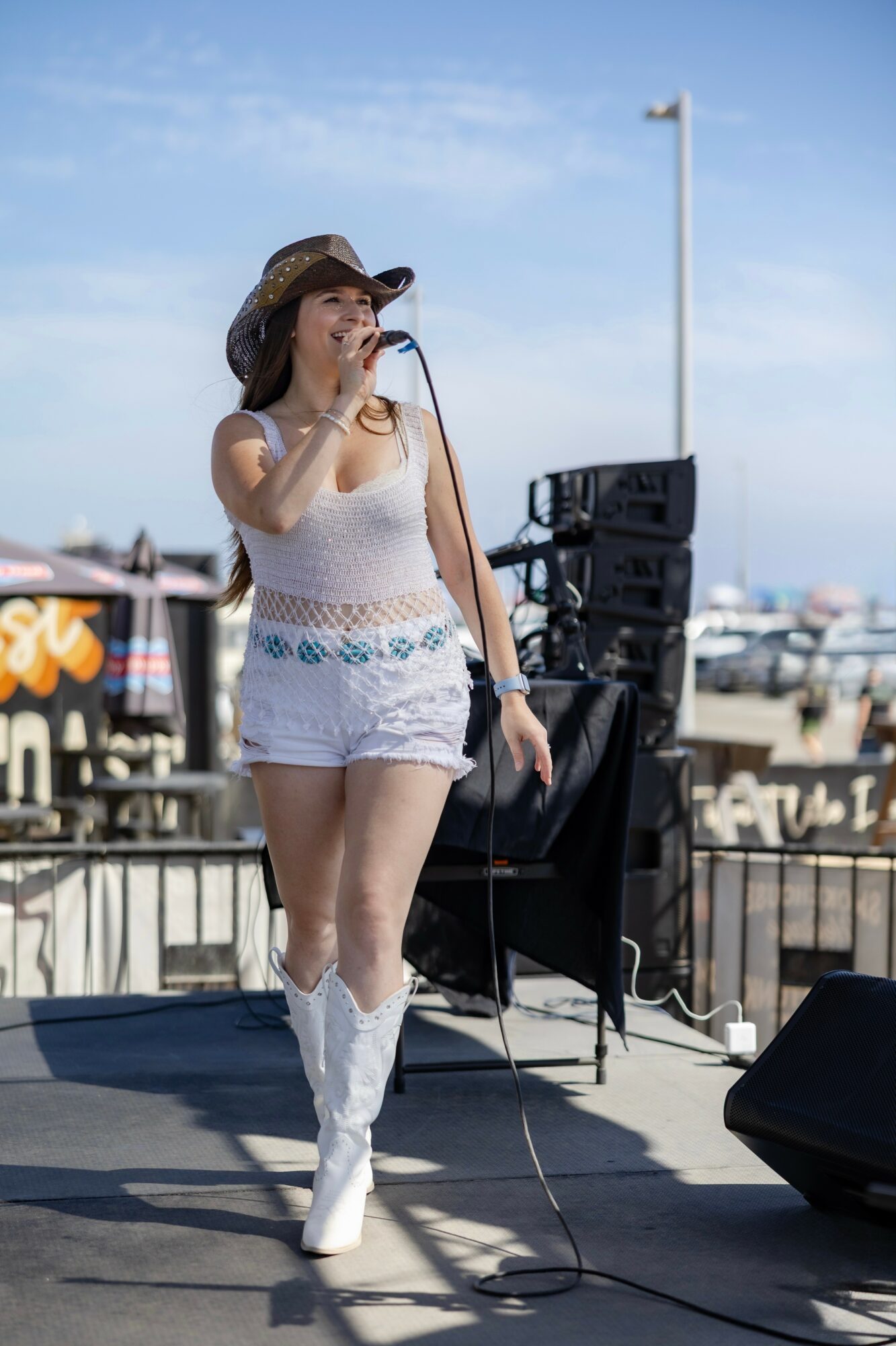 Woman singing on stage outdoors with boats in background, wearing a hat, white outfit, and boots.