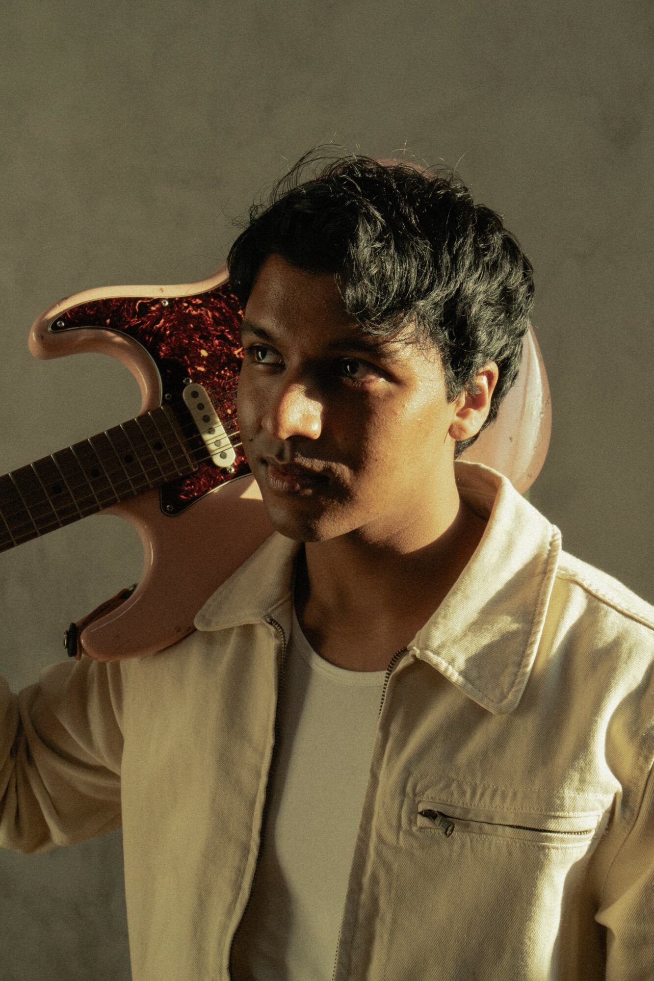 Young man with dark hair holding a guitar over shoulder, wearing a light jacket and white shirt, against a plain background.