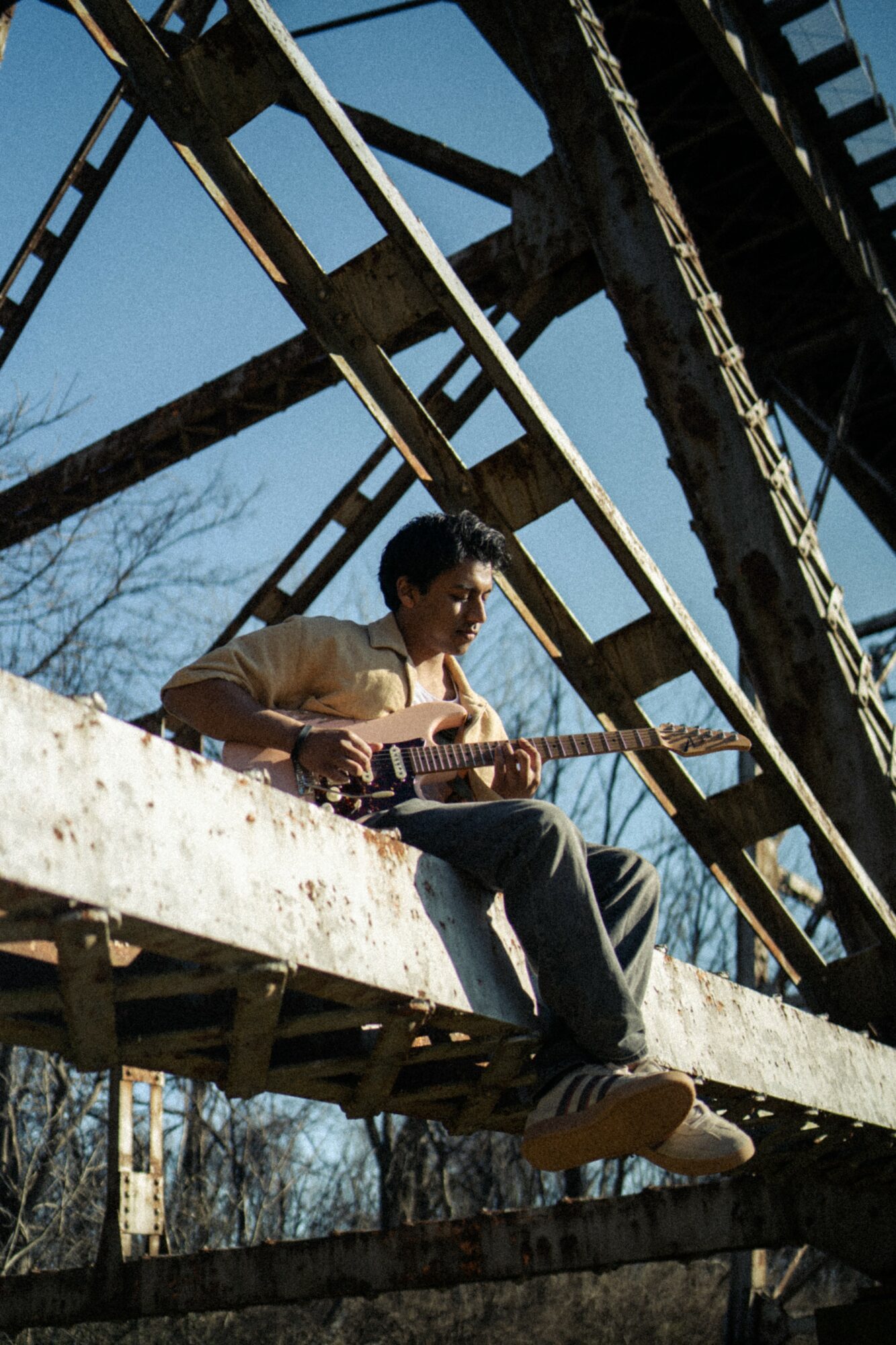 Person sitting on a metal structure playing guitar outdoors, with trees and blue sky in background.
