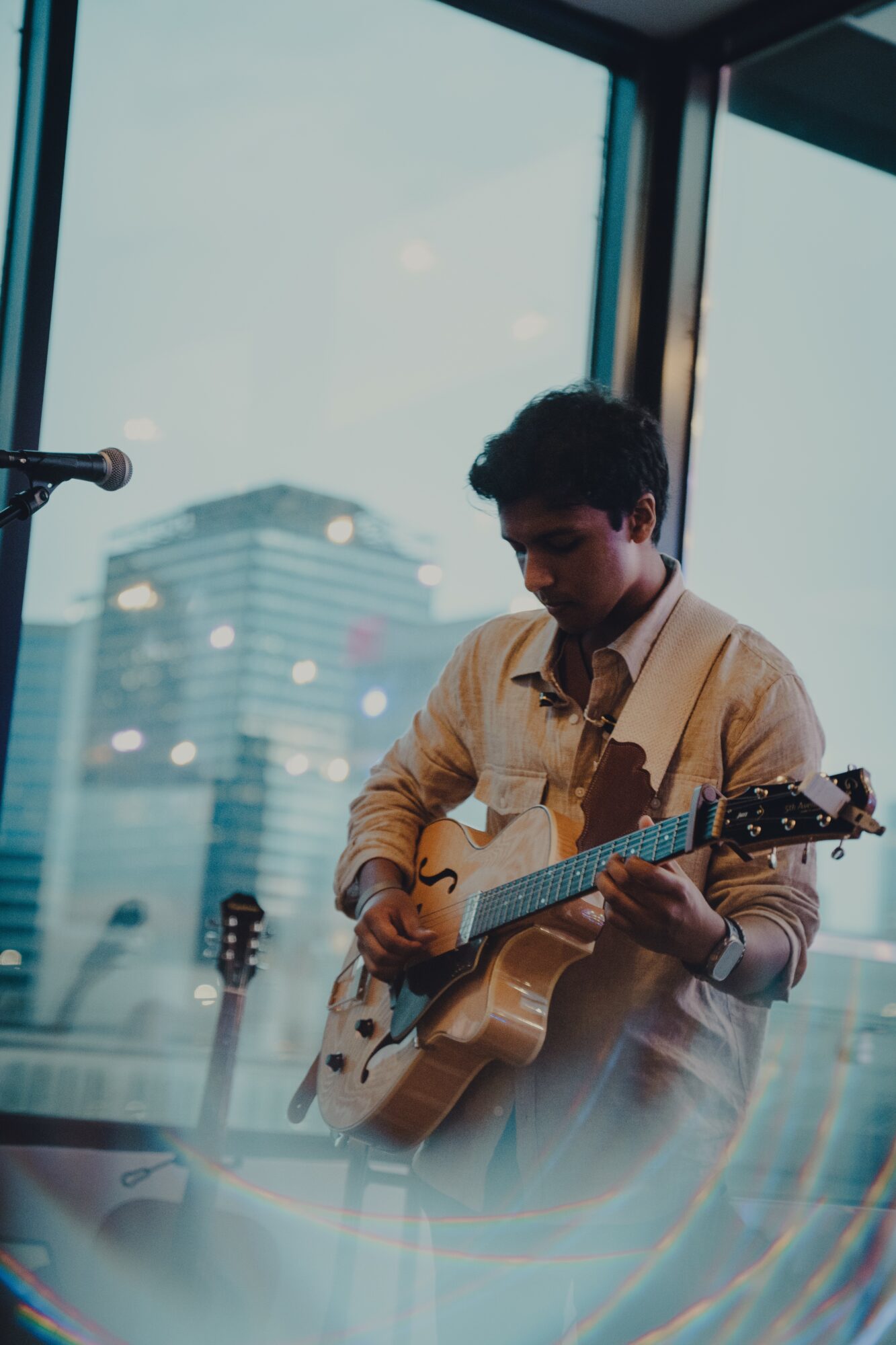 Person playing an acoustic guitar indoors with city buildings visible through large windows behind them.