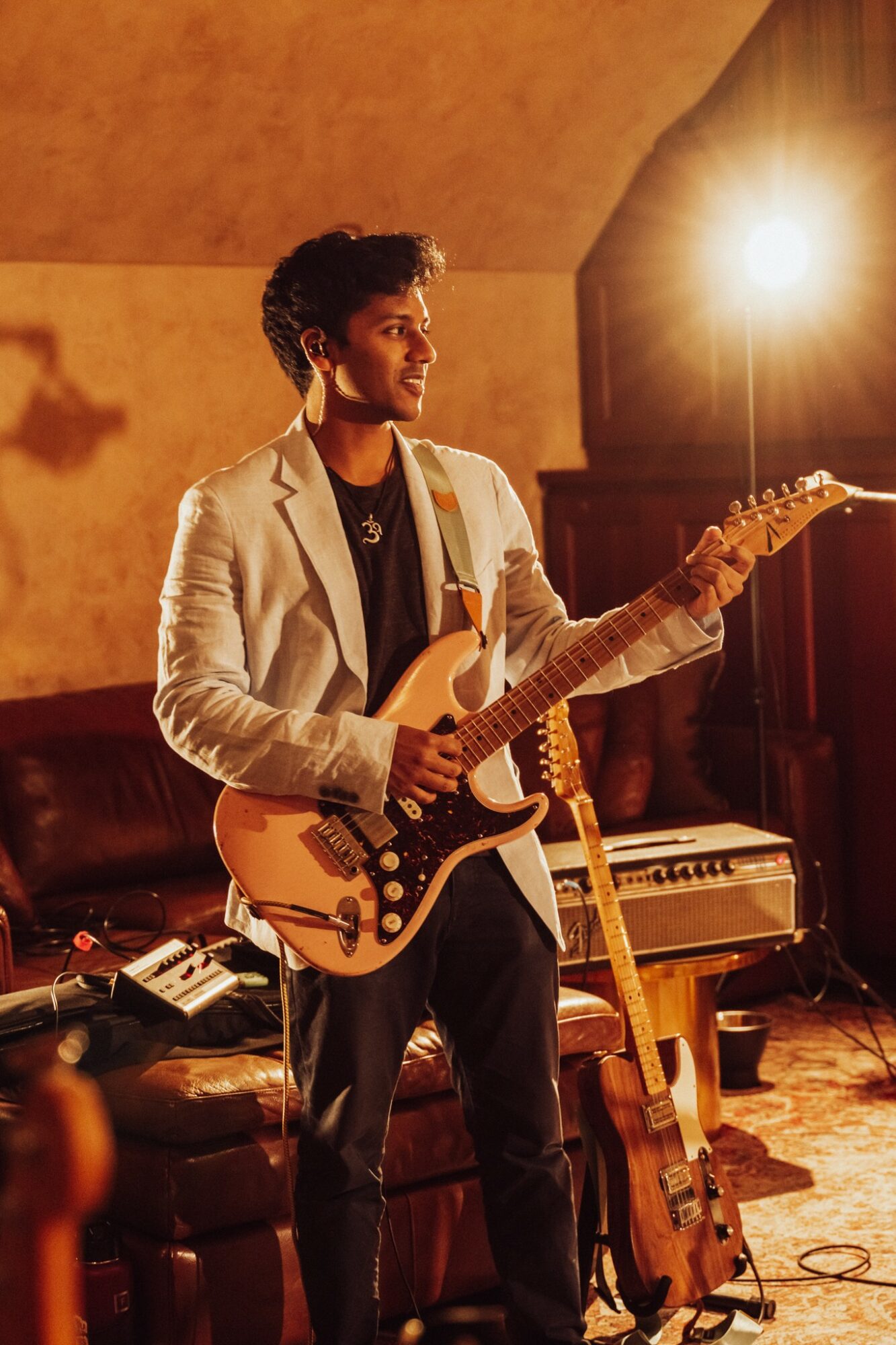 Young man playing an electric guitar on stage with warm lighting and musical equipment around him.
