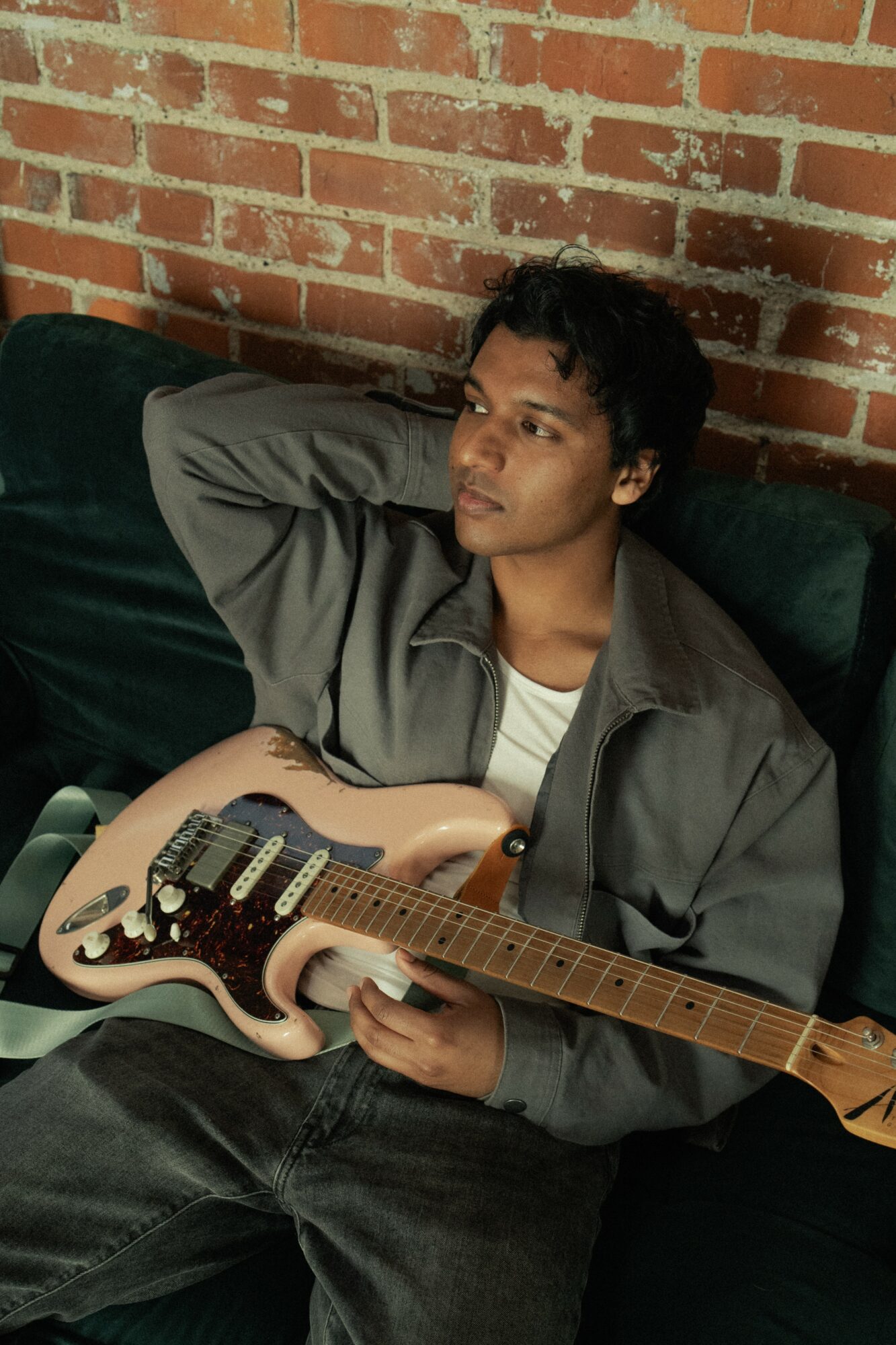 Person sitting on a dark sofa with a brick wall background, holding an electric guitar, looking to the side.
