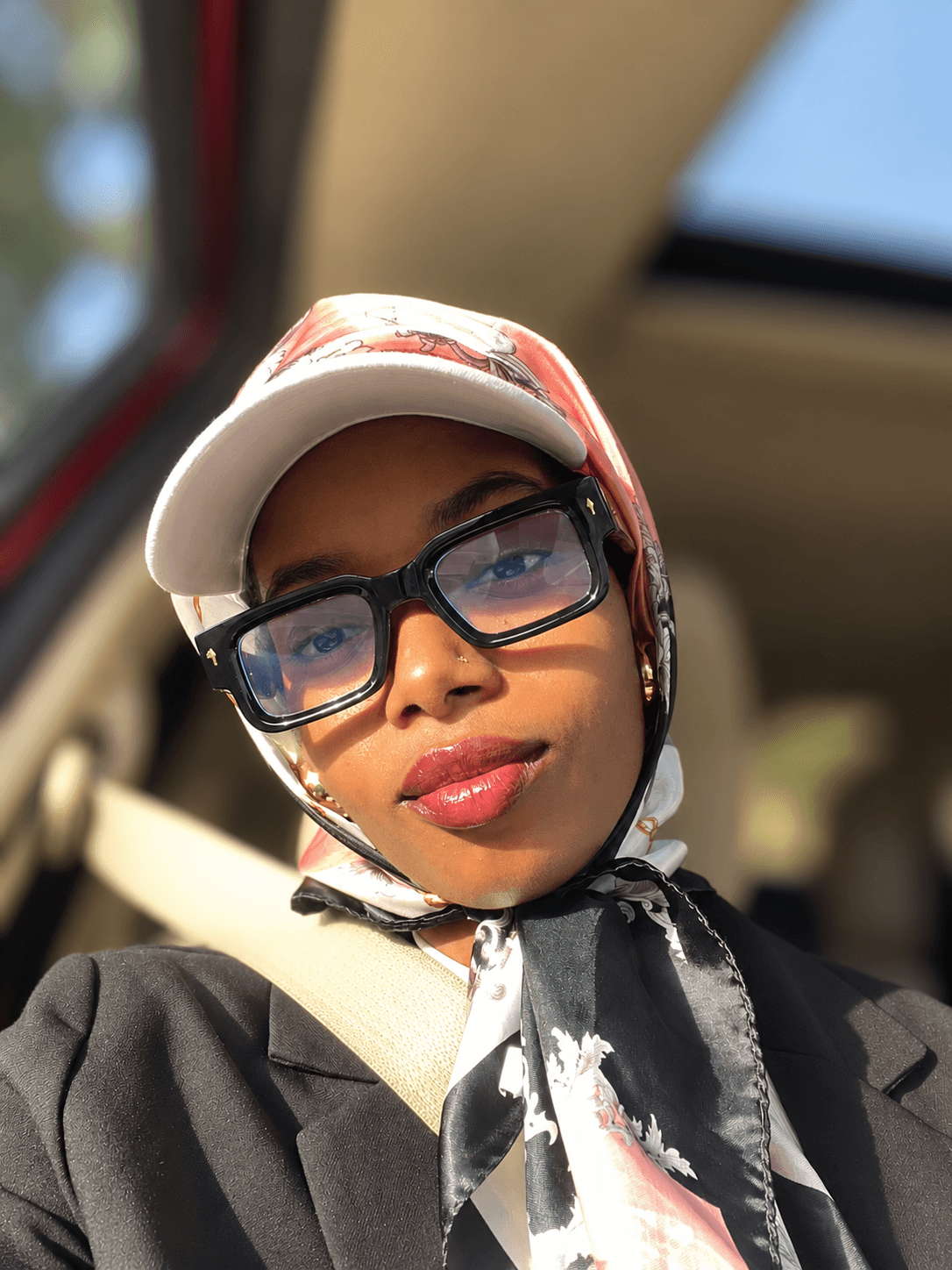 Young woman wearing glasses, a white cap, and a patterned scarf, sitting in a vehicle with sunlight on her face.