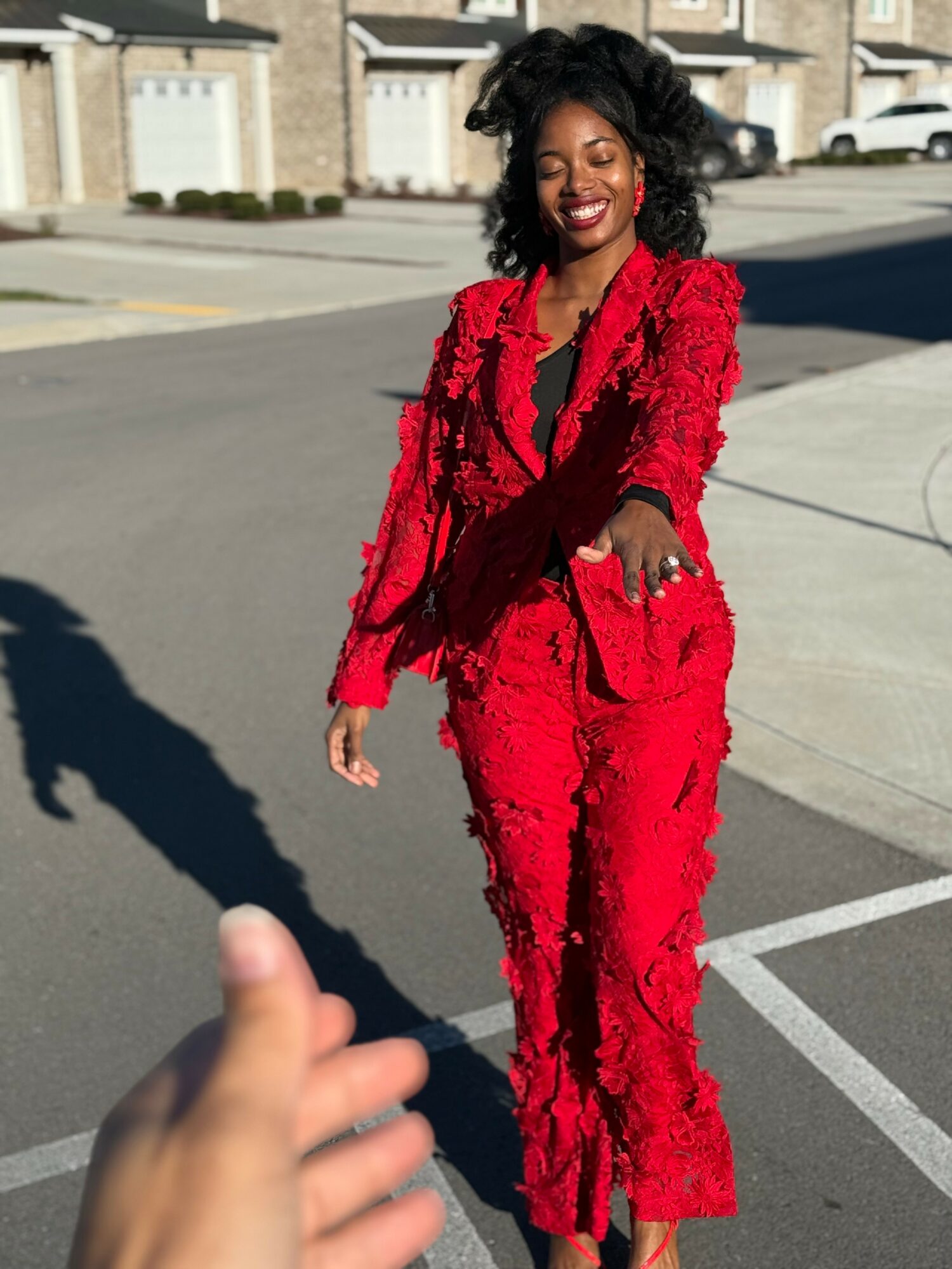 Woman in red suit smiling, standing outdoors on a sunny street, with houses in the background.