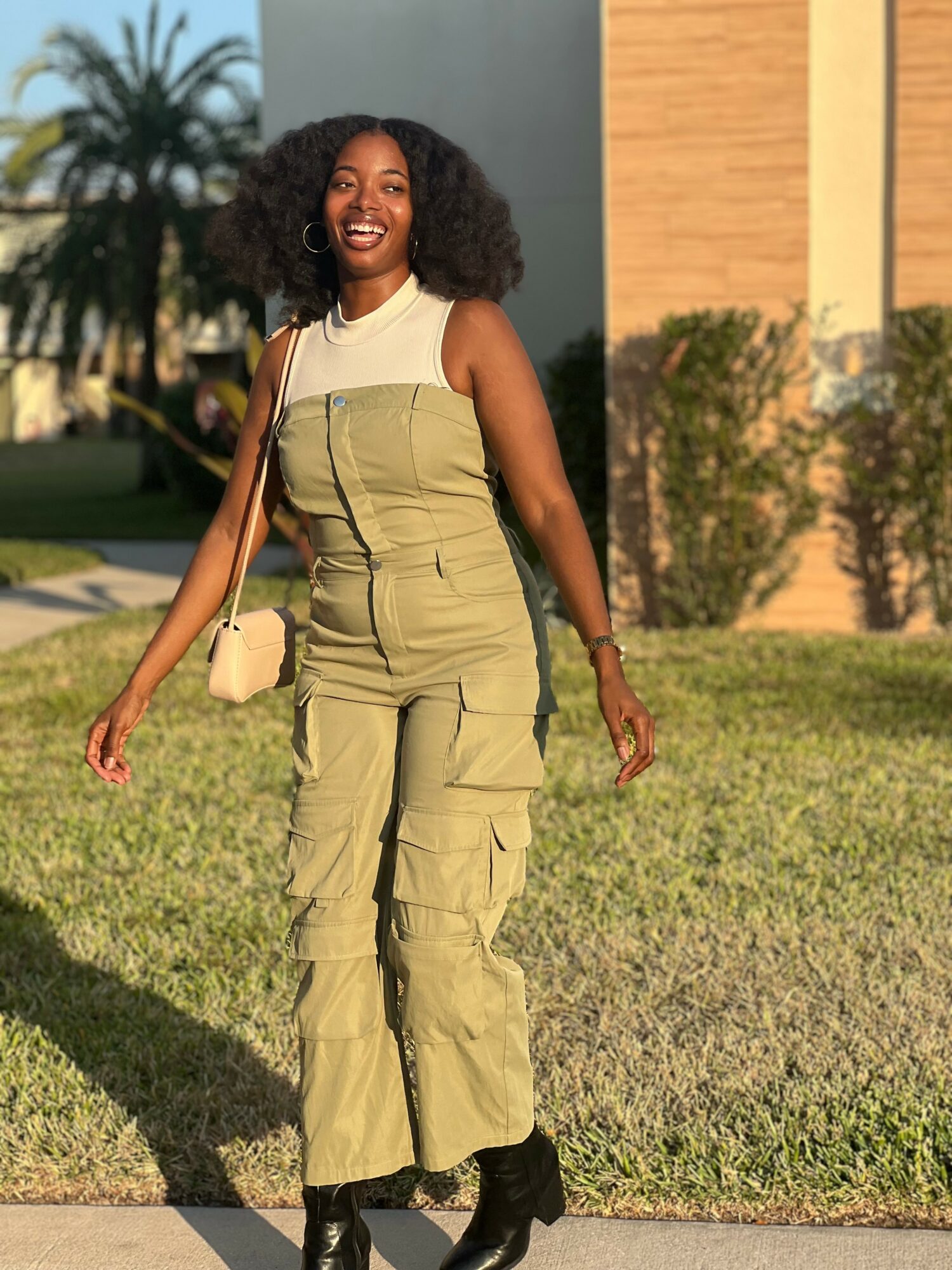 Woman with curly hair smiling outdoors, wearing a green jumpsuit and black boots, holding a beige bag.