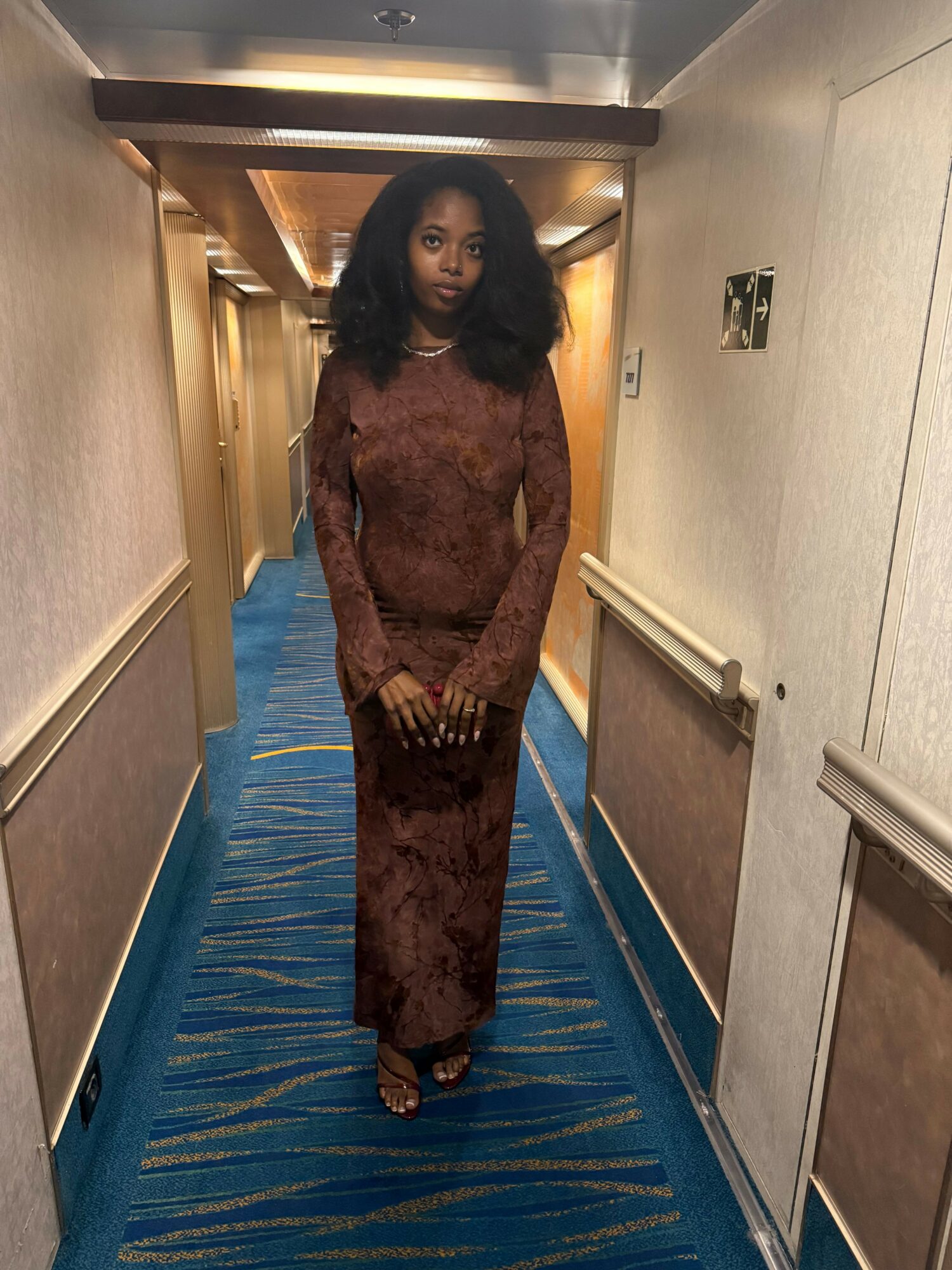 Woman in brown patterned outfit standing in hallway with blue carpet and beige walls.