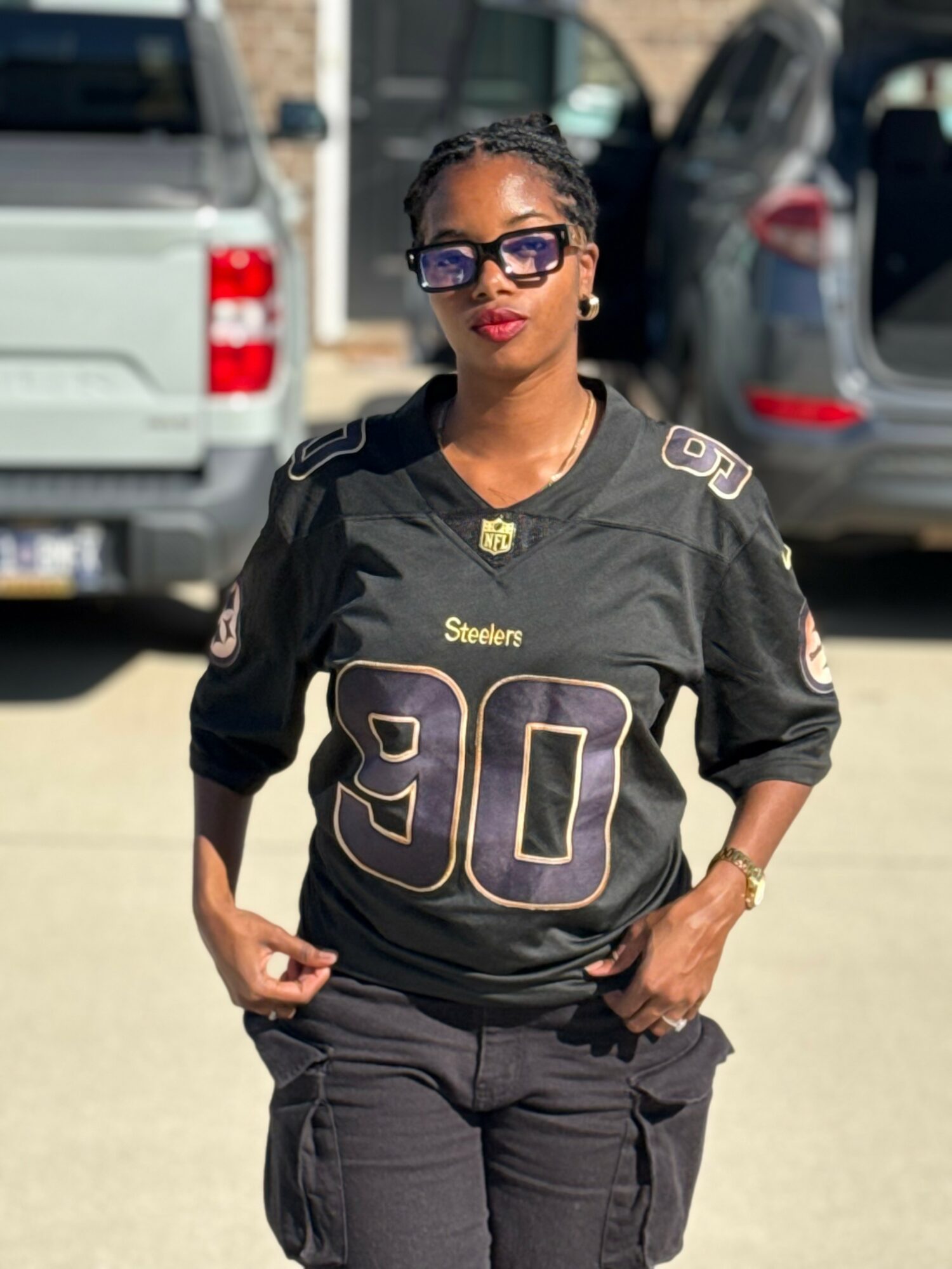 Woman wearing sunglasses and a football jersey with the number 90, standing outdoors with cars in the background.